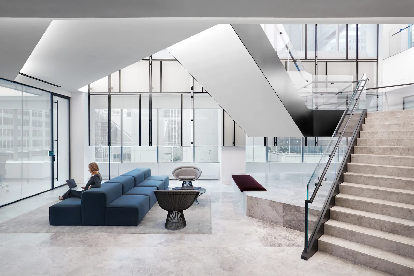 Modern office interior with a sculptural staircase, glass railings, blue sectional sofa, and floor-to-ceiling windows overlooking a cityscape