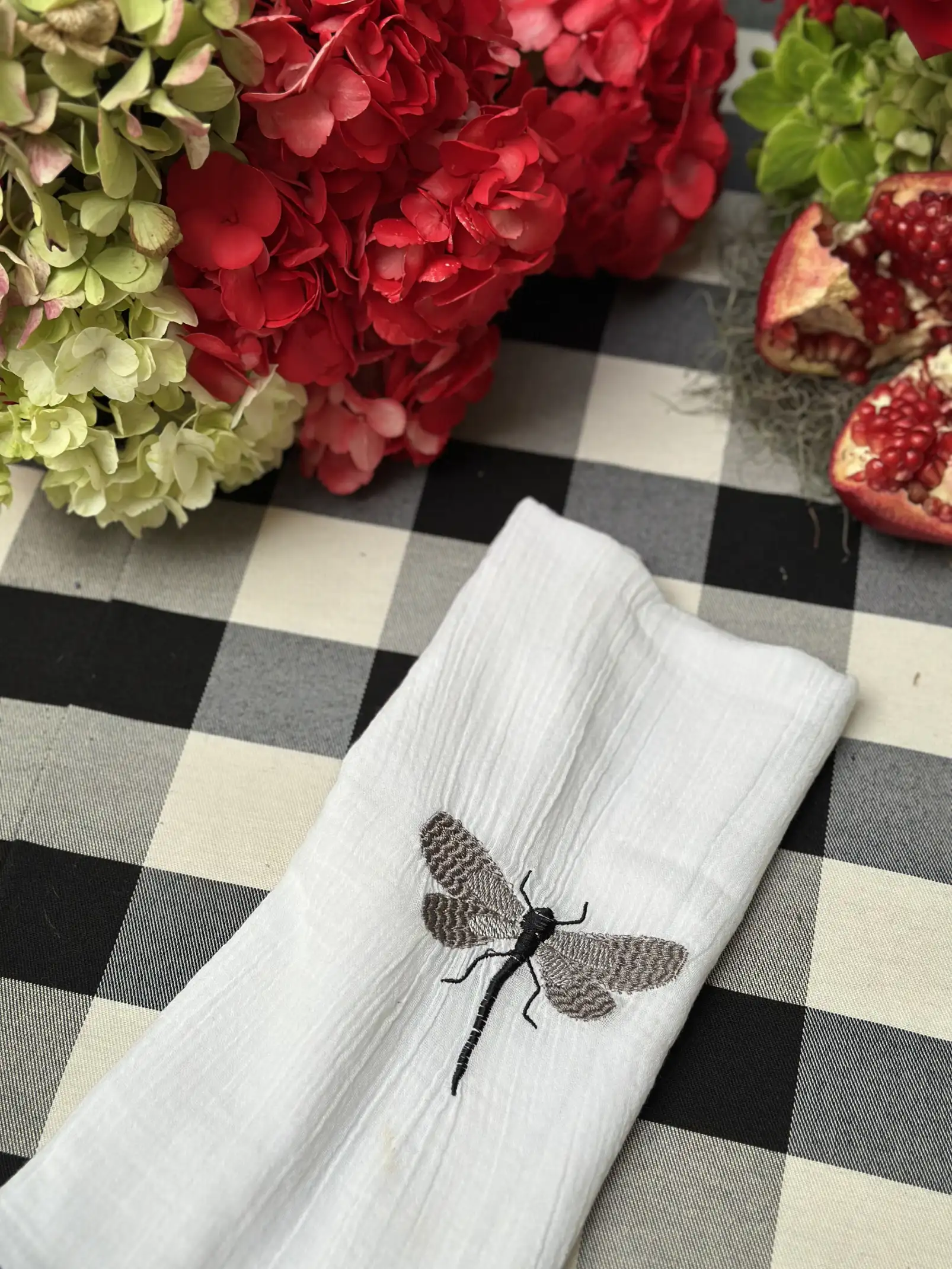 Detalle de mesa con servilleta blanca bordada con libélula, hortensias rojas y granada abierta sobre mantel a cuadros, evocando elegancia natural.