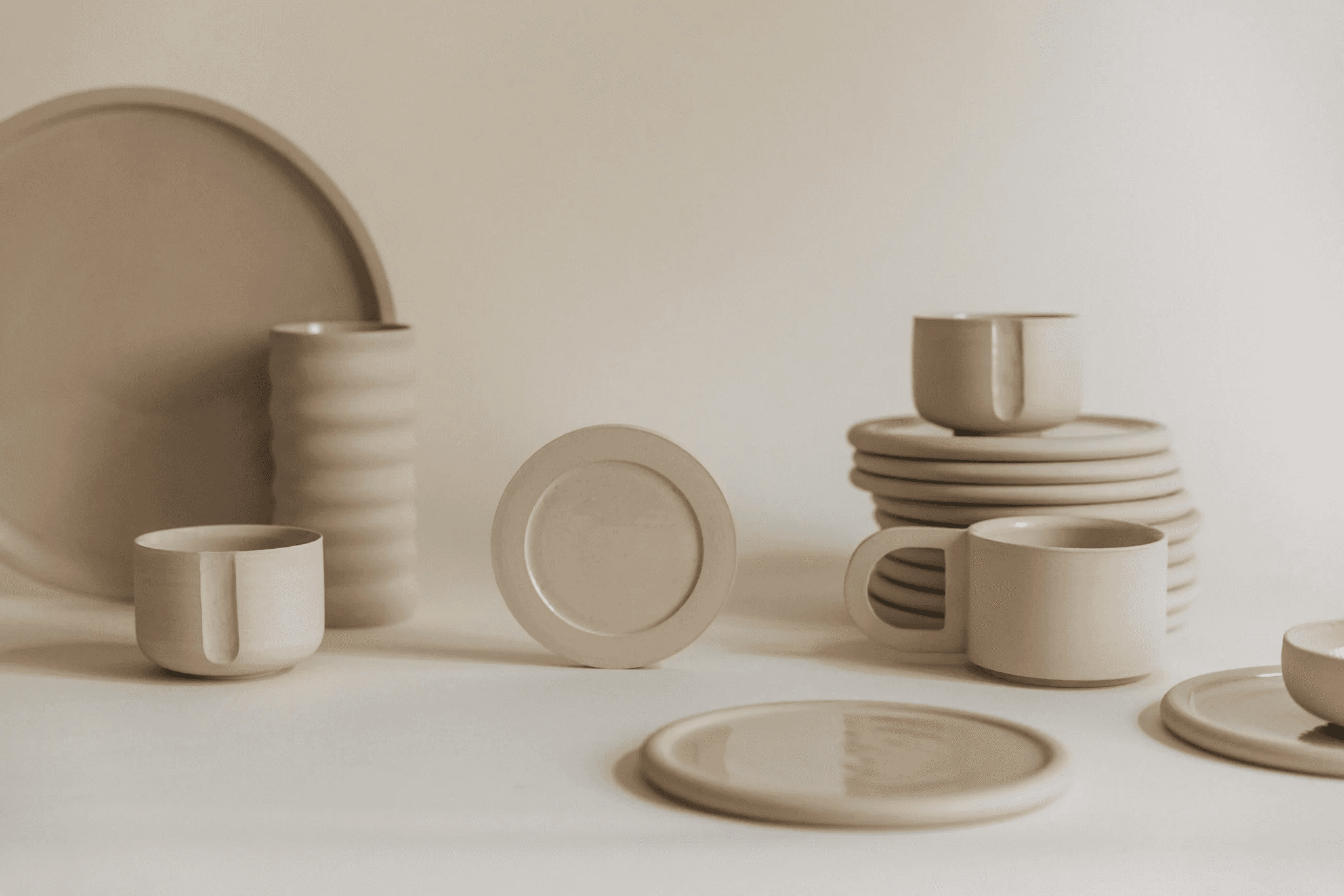 This image features an assortment of minimalist, beige ceramic dinnerware, including plates, bowls, and mugs, arranged on a cream background, embodying a modern and elegant dining aesthetic.