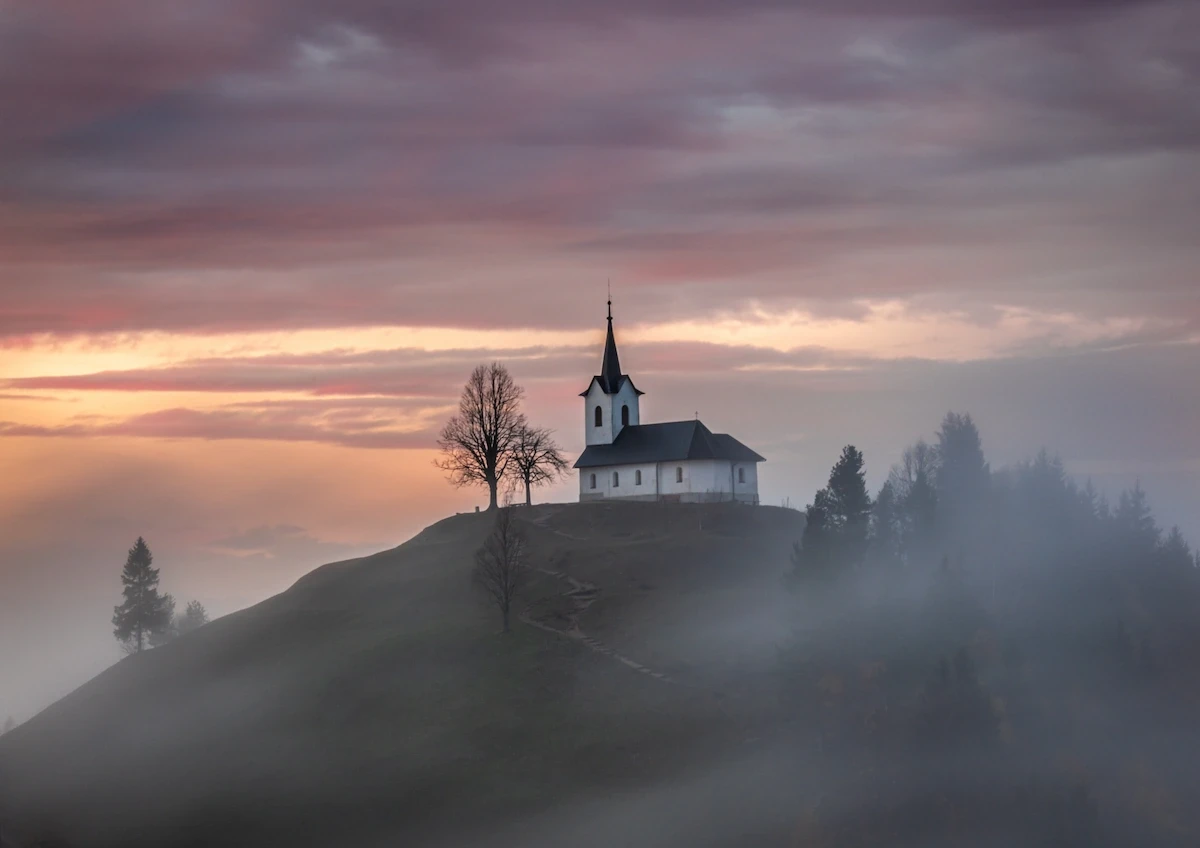 The iconic hilltop church of Sveti Jakob in Slovenia, appearing as an island above a dense layer of low-lying fog during a pastel sunrise.