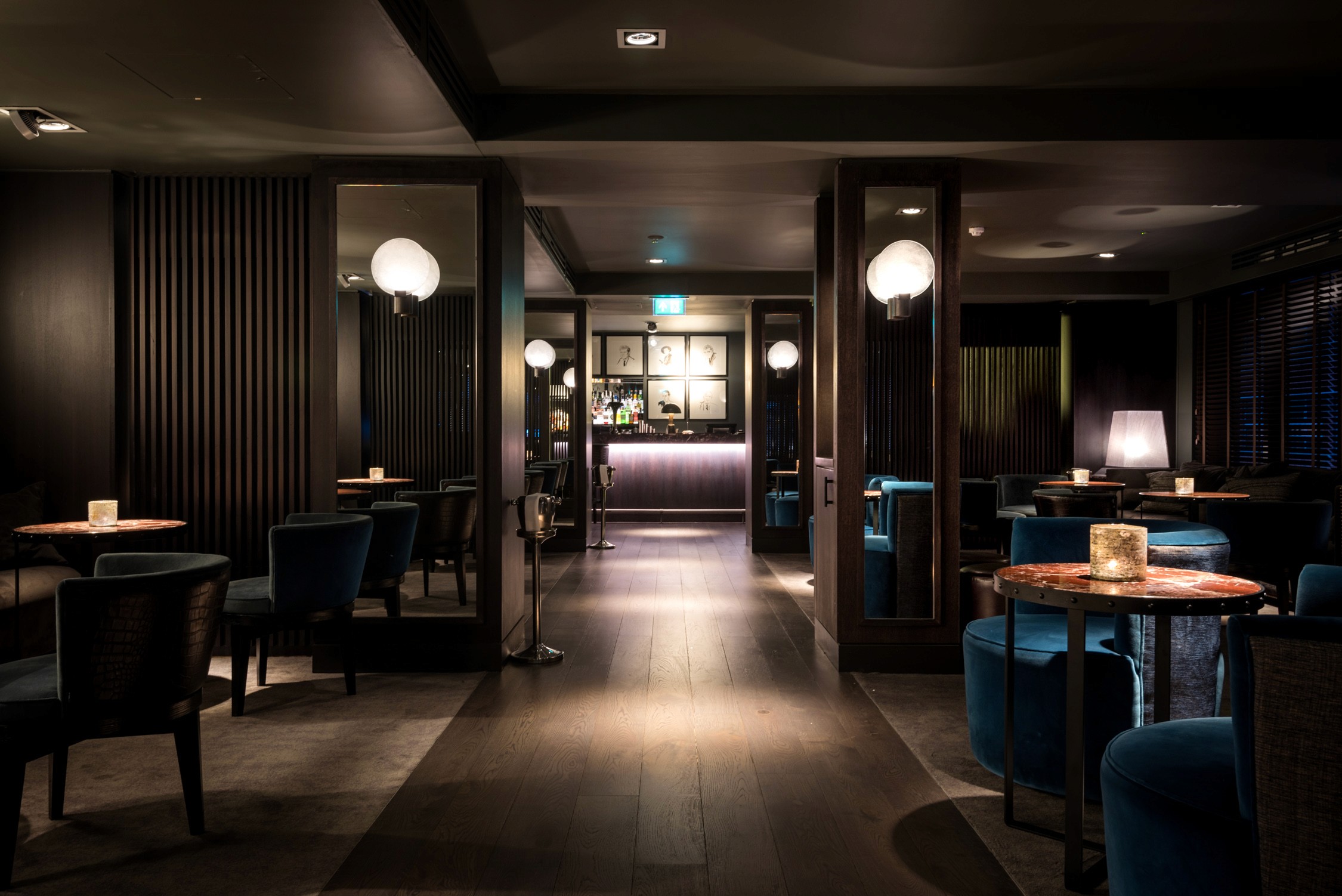 A dimly lit lounge with dark wood paneling, round tables with candles, blue velvet chairs, and glowing globe pendants.