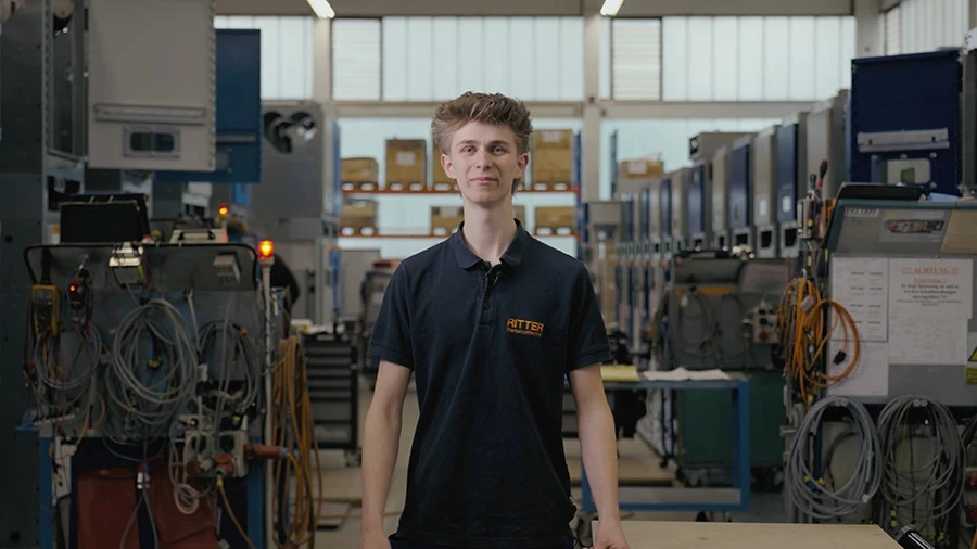 Auszubildender Shawn von RITTER Starkstromtechnik steht in der Ausbildungswerkstatt und blickt in die Kamera. Im Hintergrund sind Schaltschränke und Arbeitsplätze der Fertigung zu sehen.