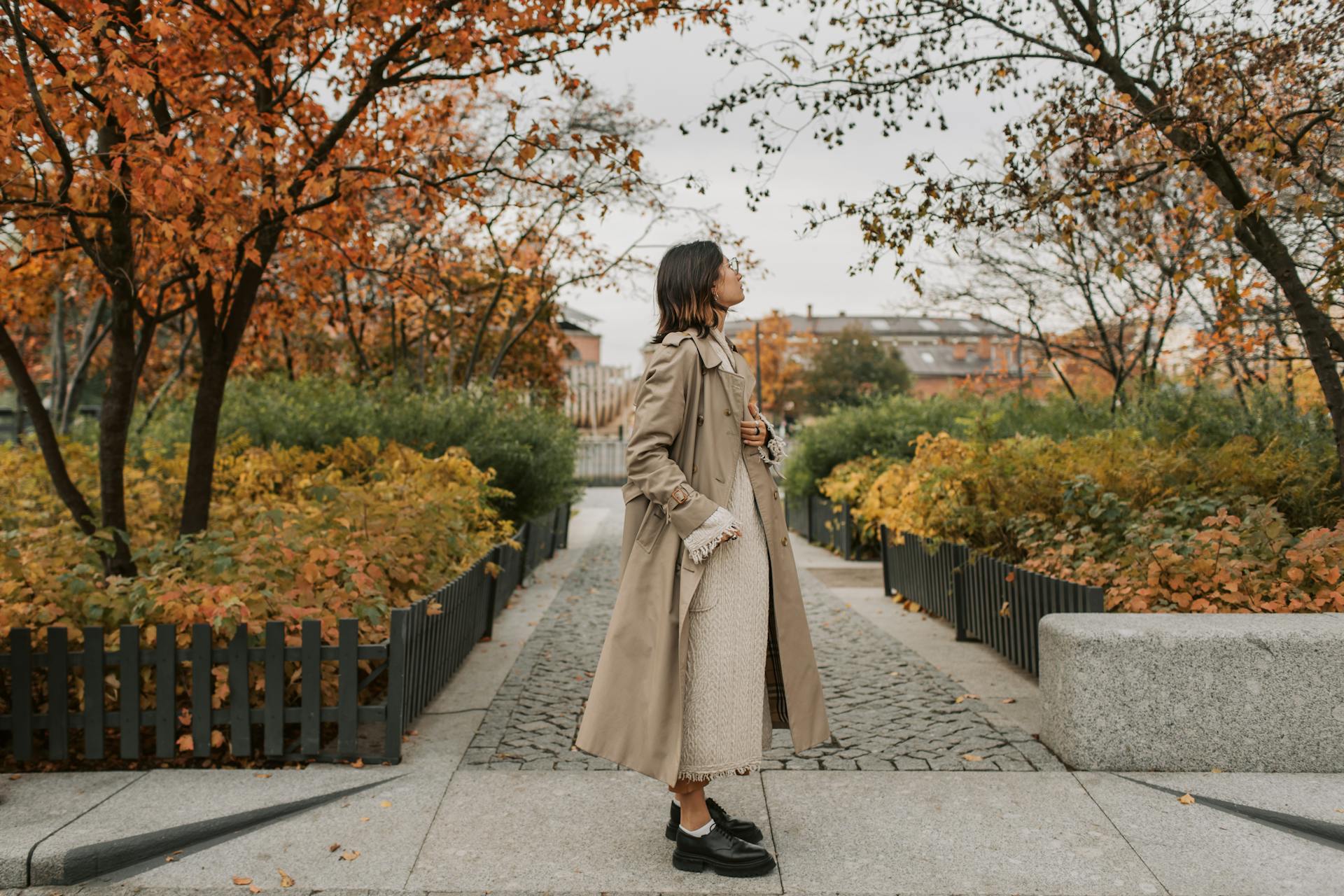 A person in a trench coat and chunky black shoes stands on a park path in autumn
