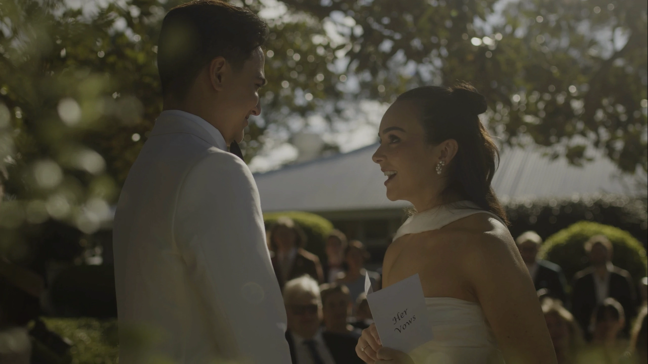 A couple stands facing each other, smiling and exchanging vows under the dappled sunlight of an outdoor garden, surrounded by greenery and seated guests in formal attire.