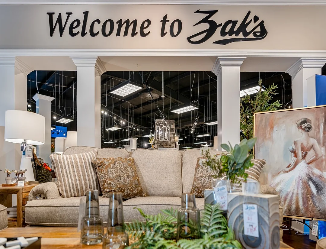 A cozy furniture store display featuring a beige sofa, decorative plants, and a "Welcome to Zak's" sign.