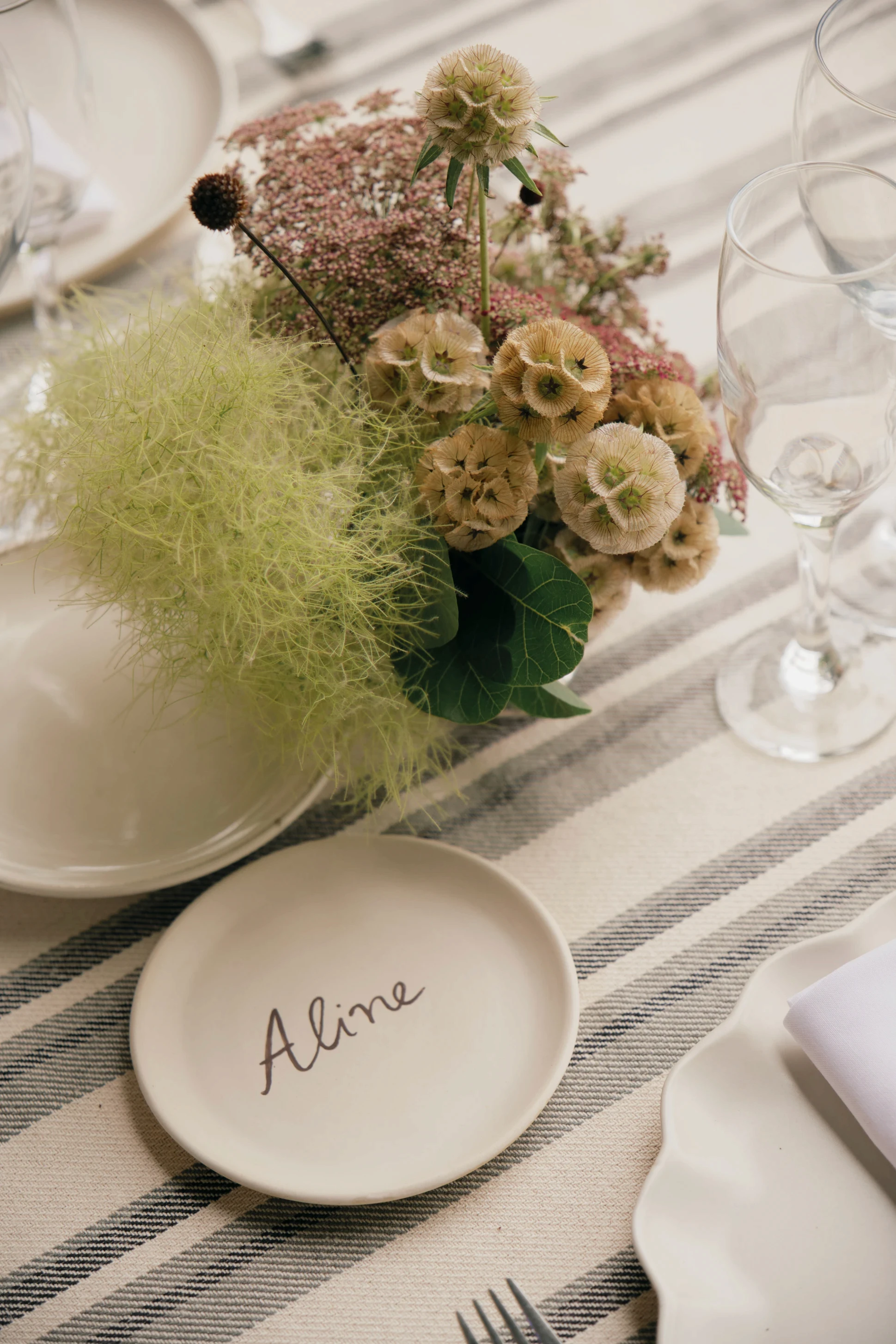 Puesta de mesa con plato de sitio personalizado con el nombre 'Aline' y centro de mesa floral etéreo.