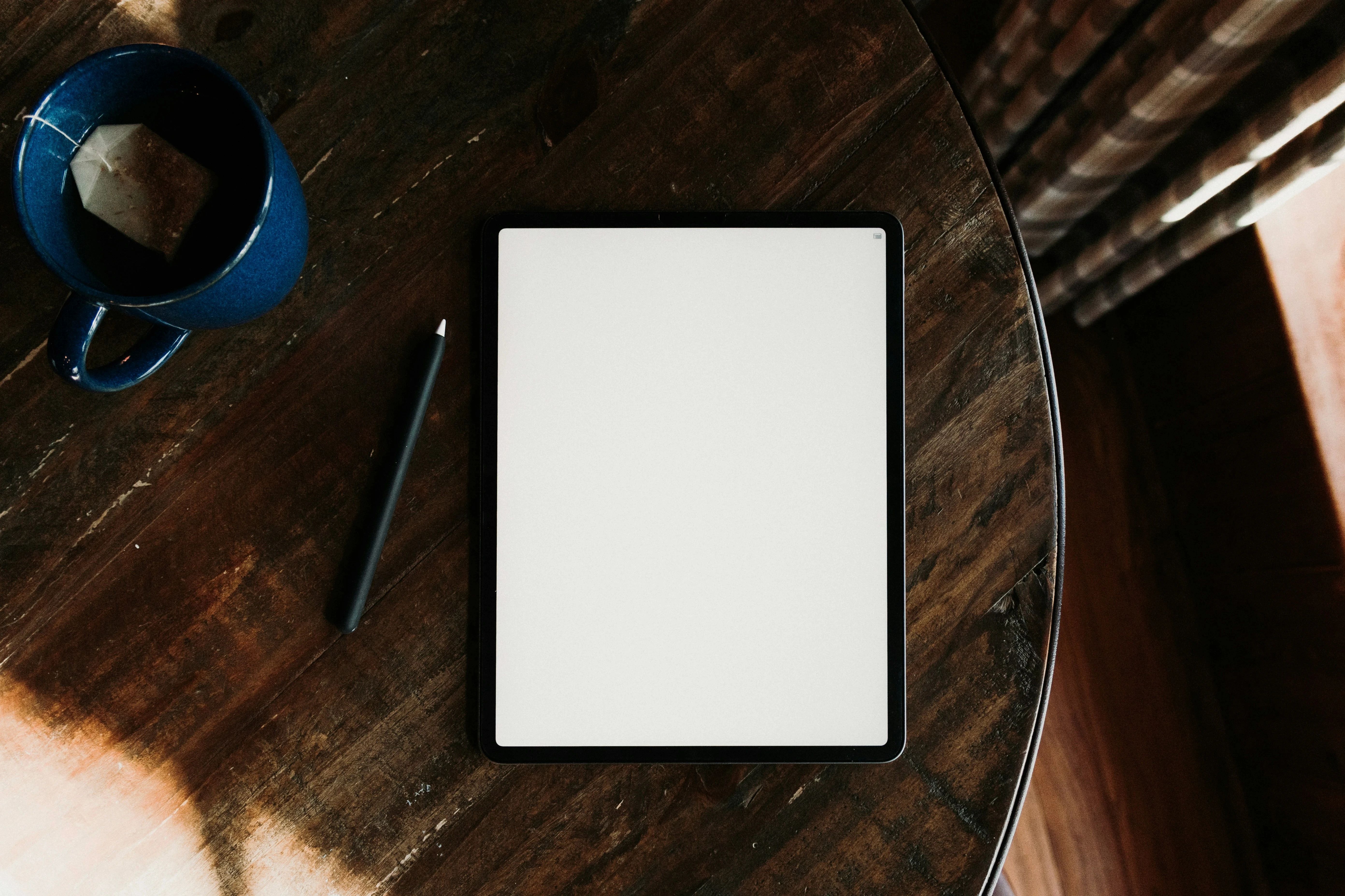 iPad with black stylus on a dark wooden table next to a blue mug of tea with a tea bag string visible.