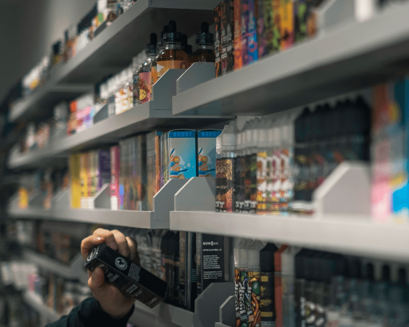 Shelves stocked with colourful vape liquids and e-cigarette products, with a hand picking up a vape device in a retail setting.