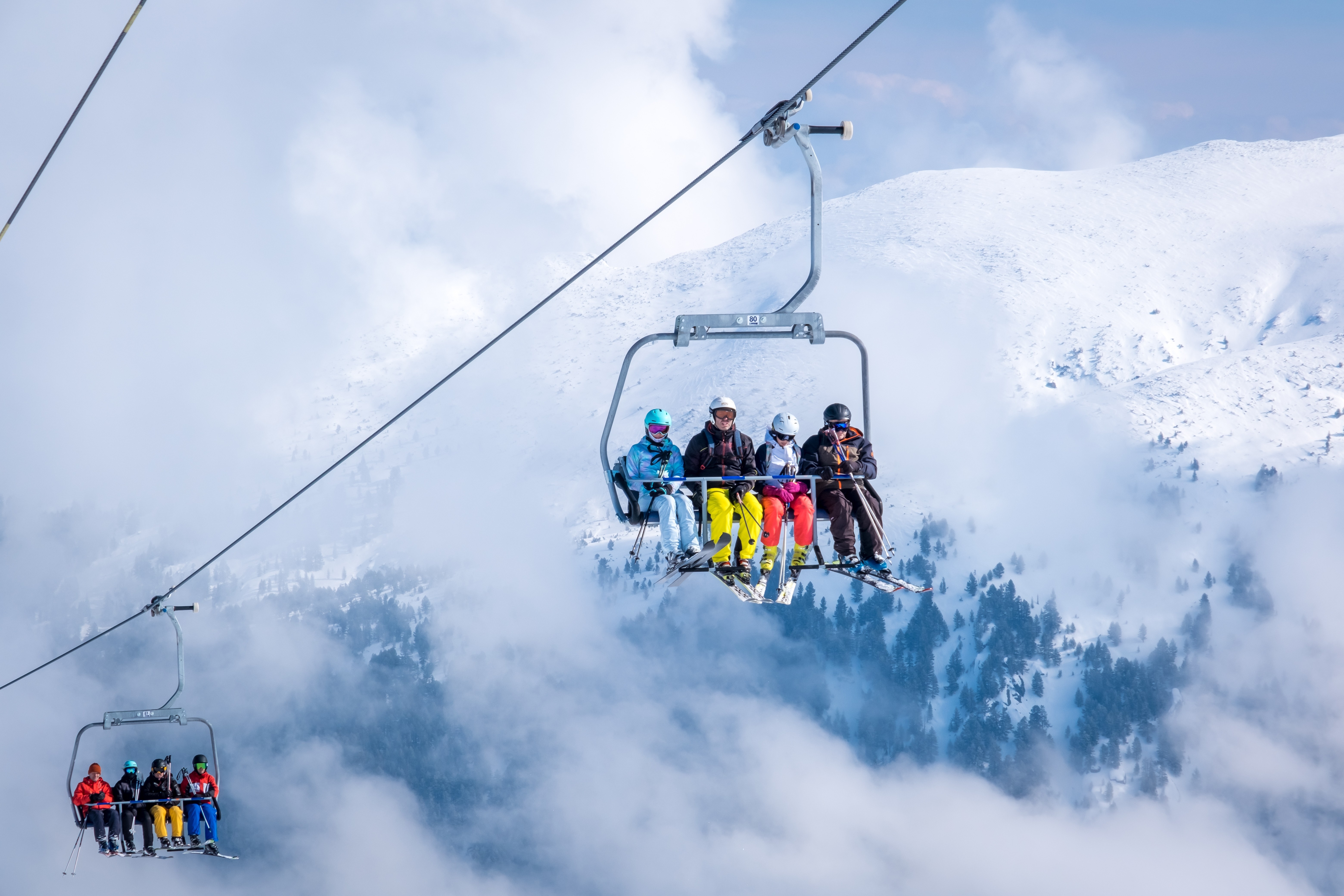 Skiers ride a chairlift above snowy mountains and drifting clouds on a bright winter day.