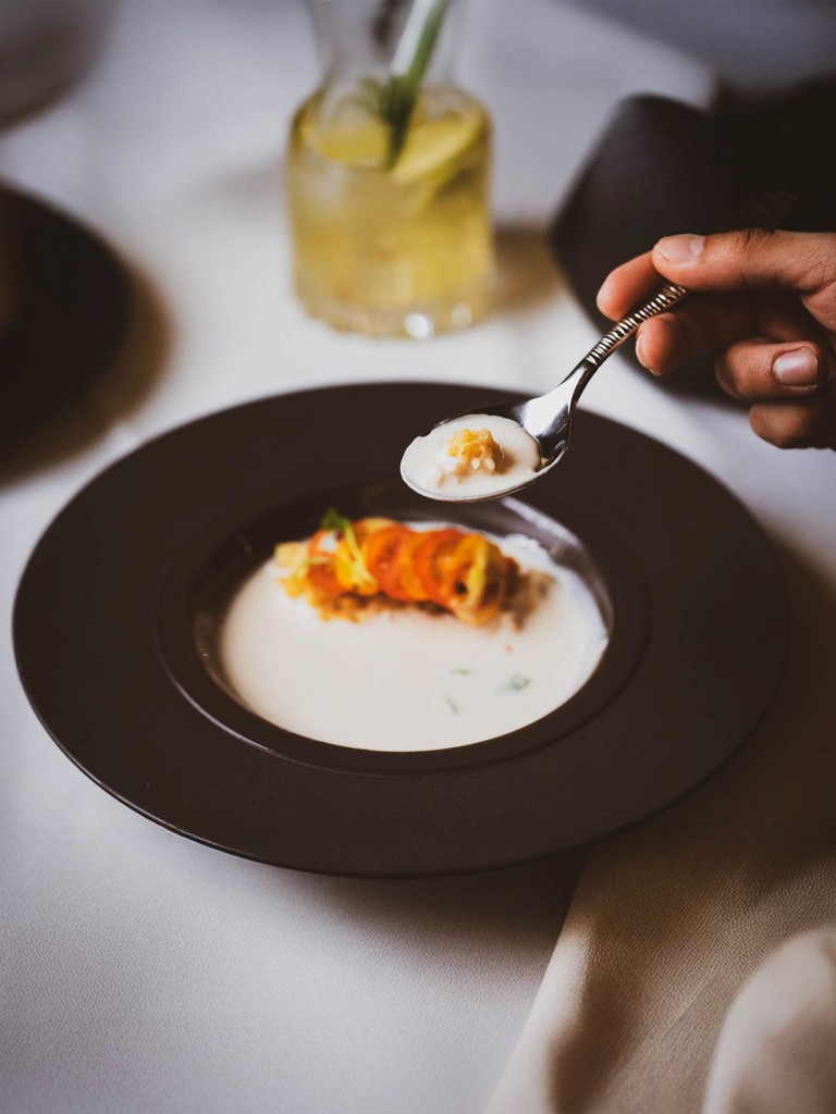 A person uses a spoon to sample a creamy soup with an orange garnish in a dark-colored bowl.