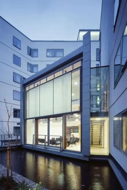 A modern villa with a sleek glass facade overlooks an infinity pool at sunset. Palm trees line the edge, with a distant city skyline aglow.