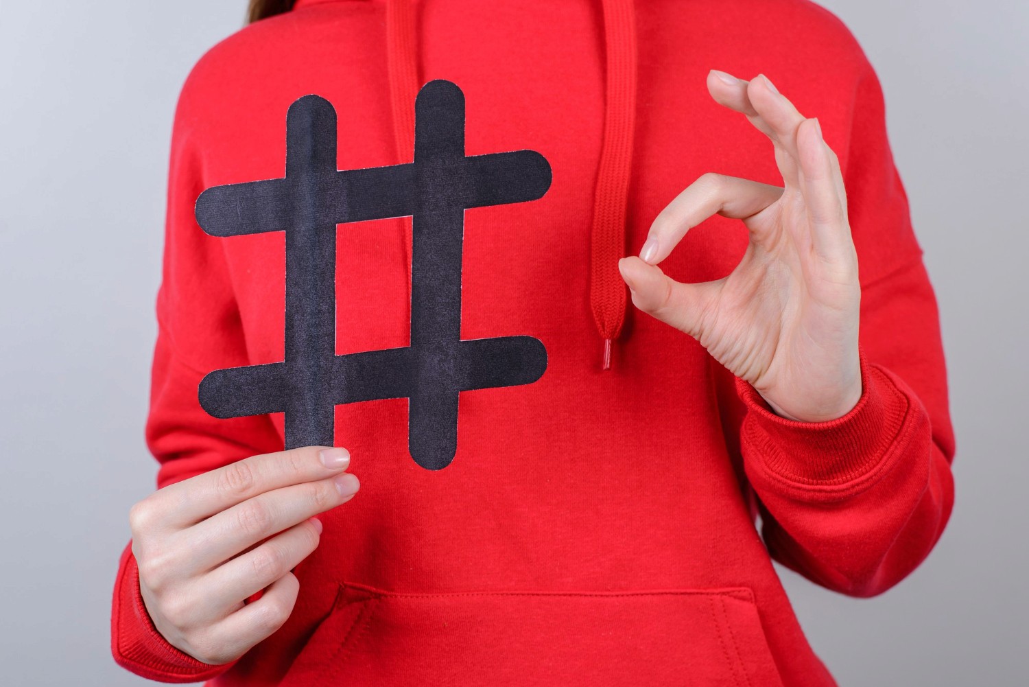 A woman in a red hoodie proudly displays a hashtag sign, symbolizing social media engagement and connection.