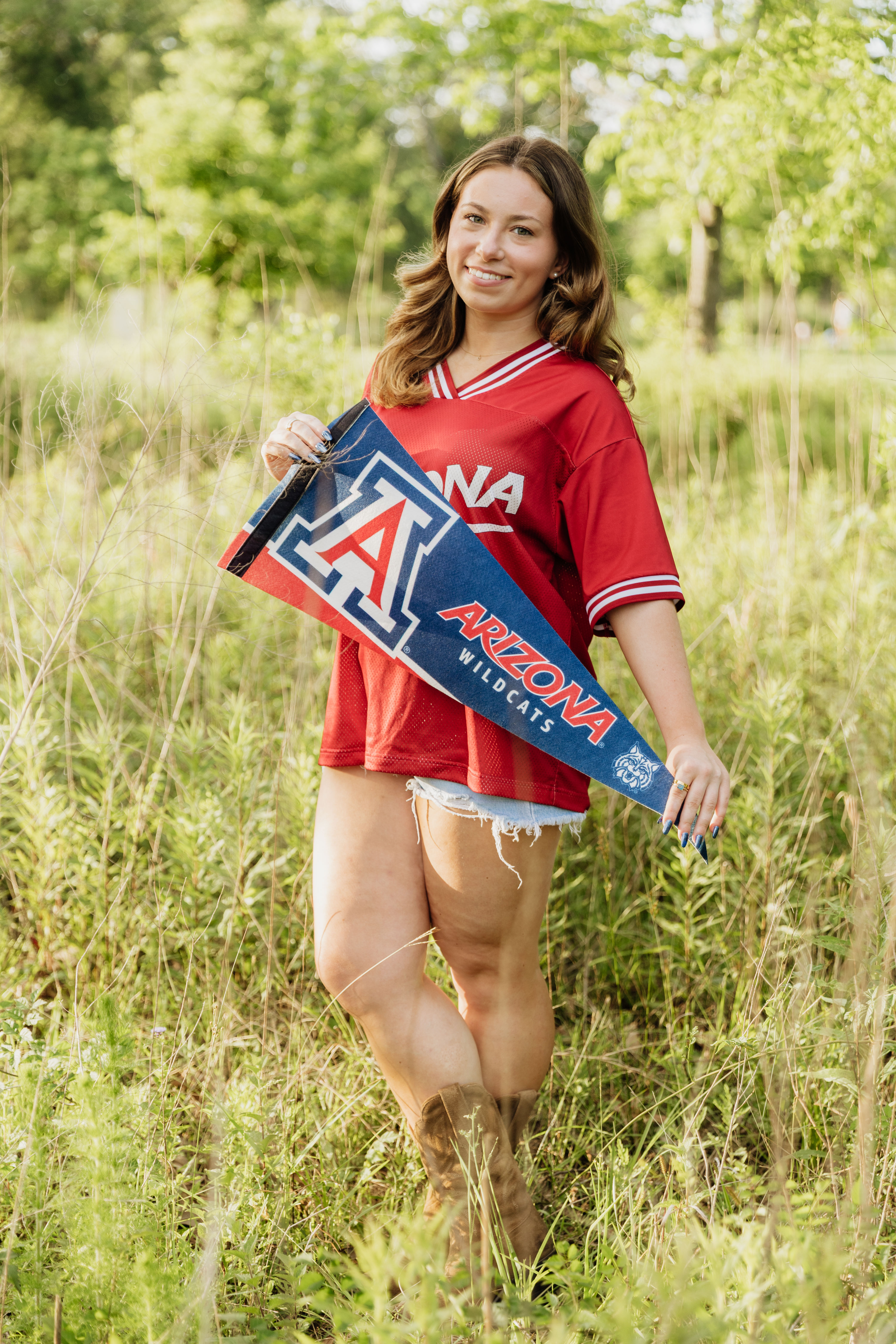 A high school senior girl smiling in a grassy field at Memorial Park in Houston, wearing a red Arizona Wildcats jersey and holding a blue University of Arizona pennant.