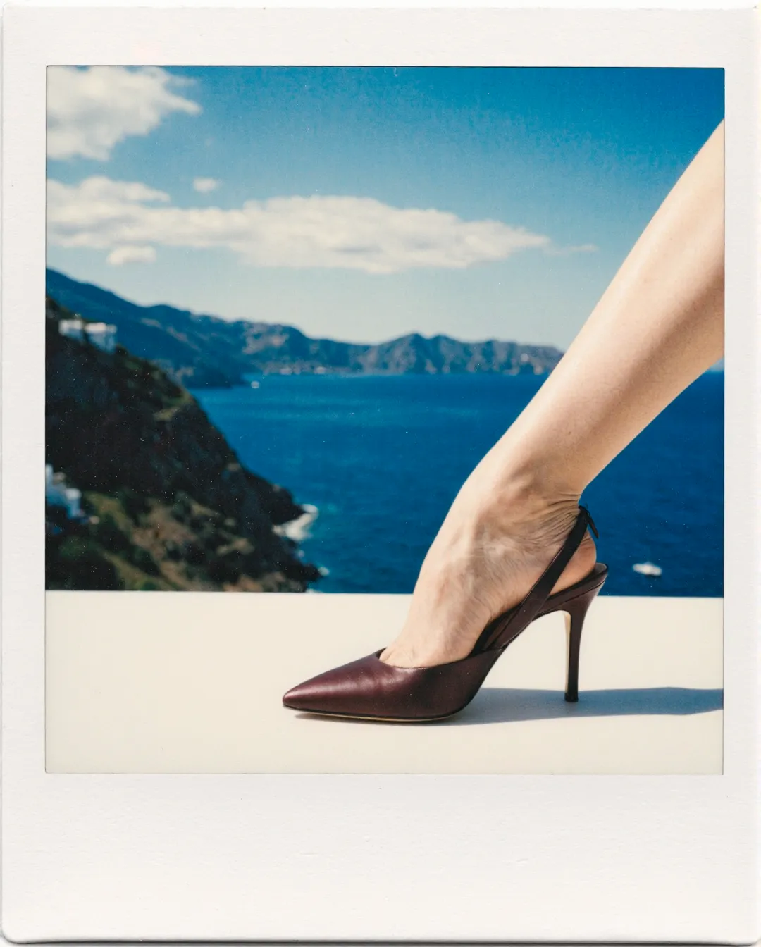 Burgundy leather stiletto slingback heel positioned on white surface with dramatic Mediterranean coastline and blue water in background