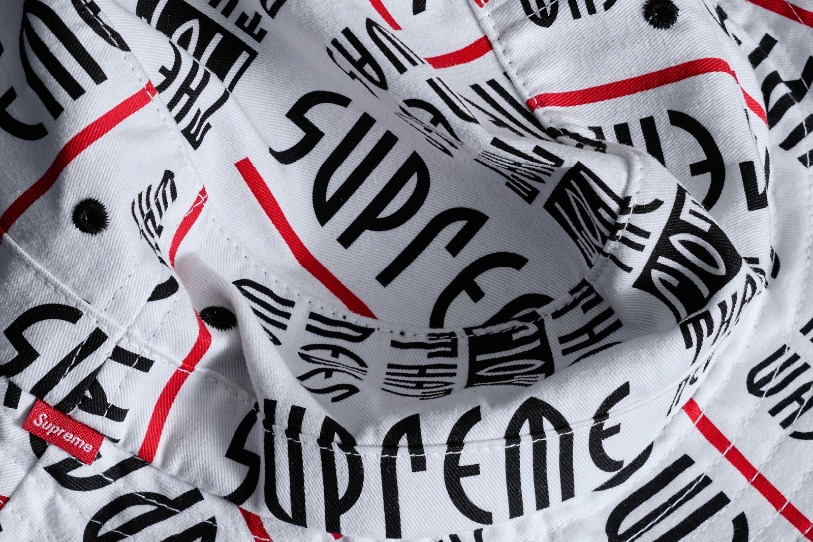 A close-up of fabric on a bucket hat featuring a repeated pattern of the word "SUPREME" in black and red against a white background.