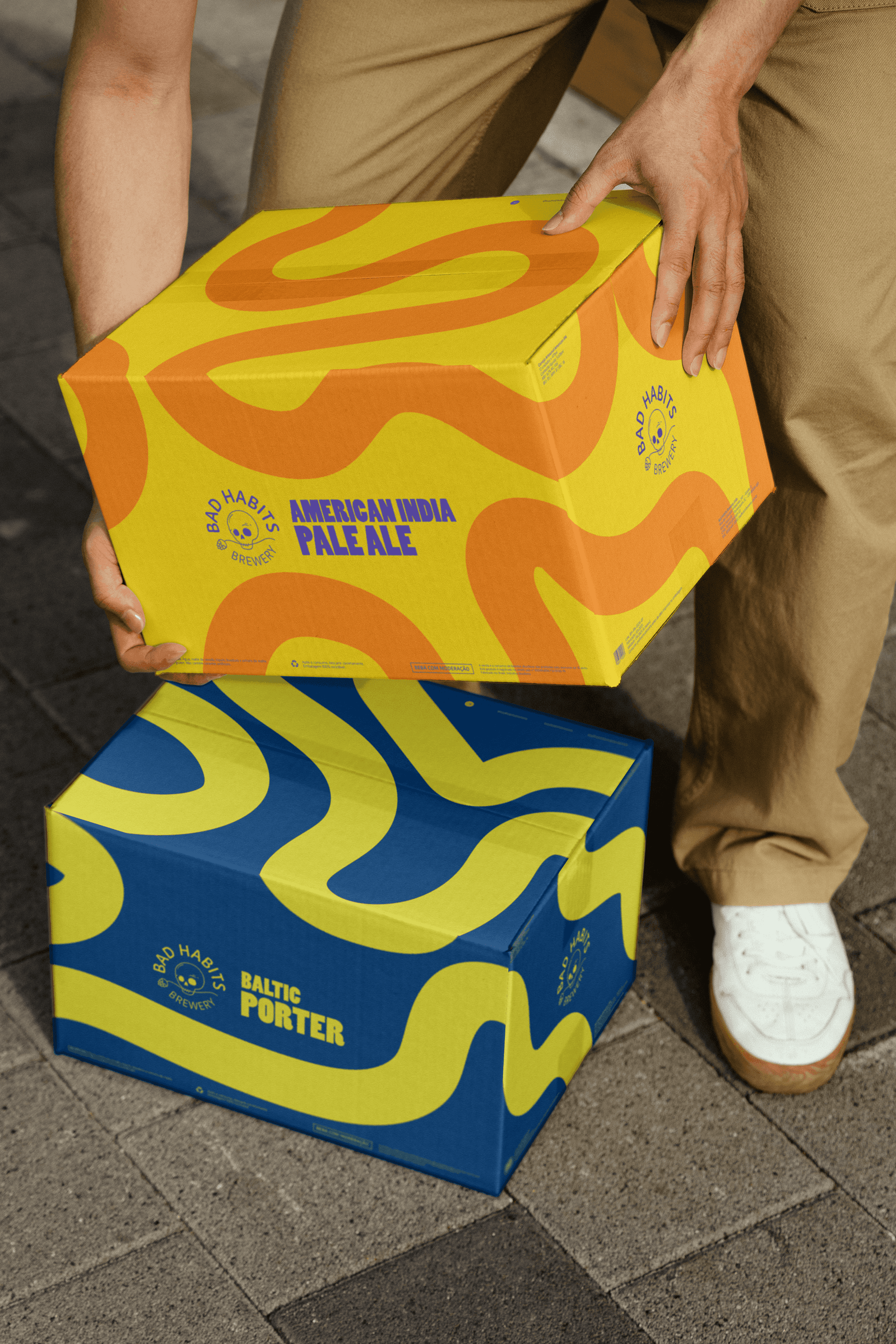 A person lifting a yellow and orange Bad Habits Brewery American India Pale Ale case box next to a blue Baltic Porter beer box on the ground.