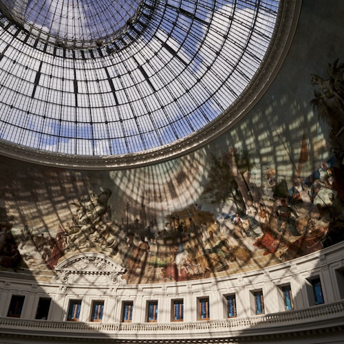 Glass-domed ceiling with intricate iron framework, casting shadows on a fresco-adorned wall in a classical interior space.
