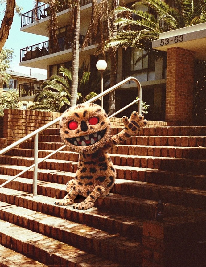 A playful character named Lampard, with fur patterned like a leopard and glowing red eyes, waves while sitting on the brick stairs outside a modern apartment building. The sunny setting features palm trees and tropical plants, giving the scene a warm, Australian vibe. Lampard’s sharp teeth and mischievous grin contrast with the relaxed outdoor backdrop.