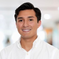 Young man with brown hair and blue eyes in an urban setting with graffiti background, wearing a dark jacket and chain necklace.