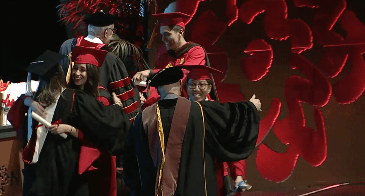 A photo of a couple of grad students and teachers, dressed in SVA color robes, that are hugging and congratulating each other.