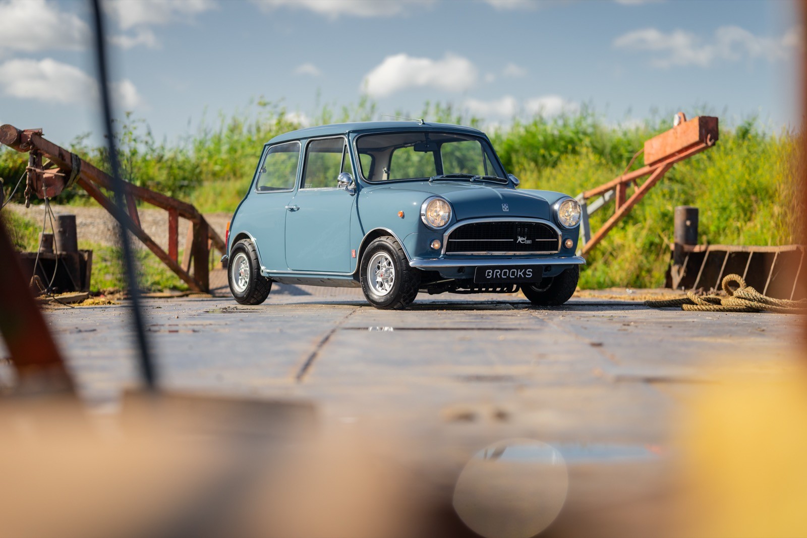 A vintage blue car parked on a road surrounded by greenery under a cloudy sky.