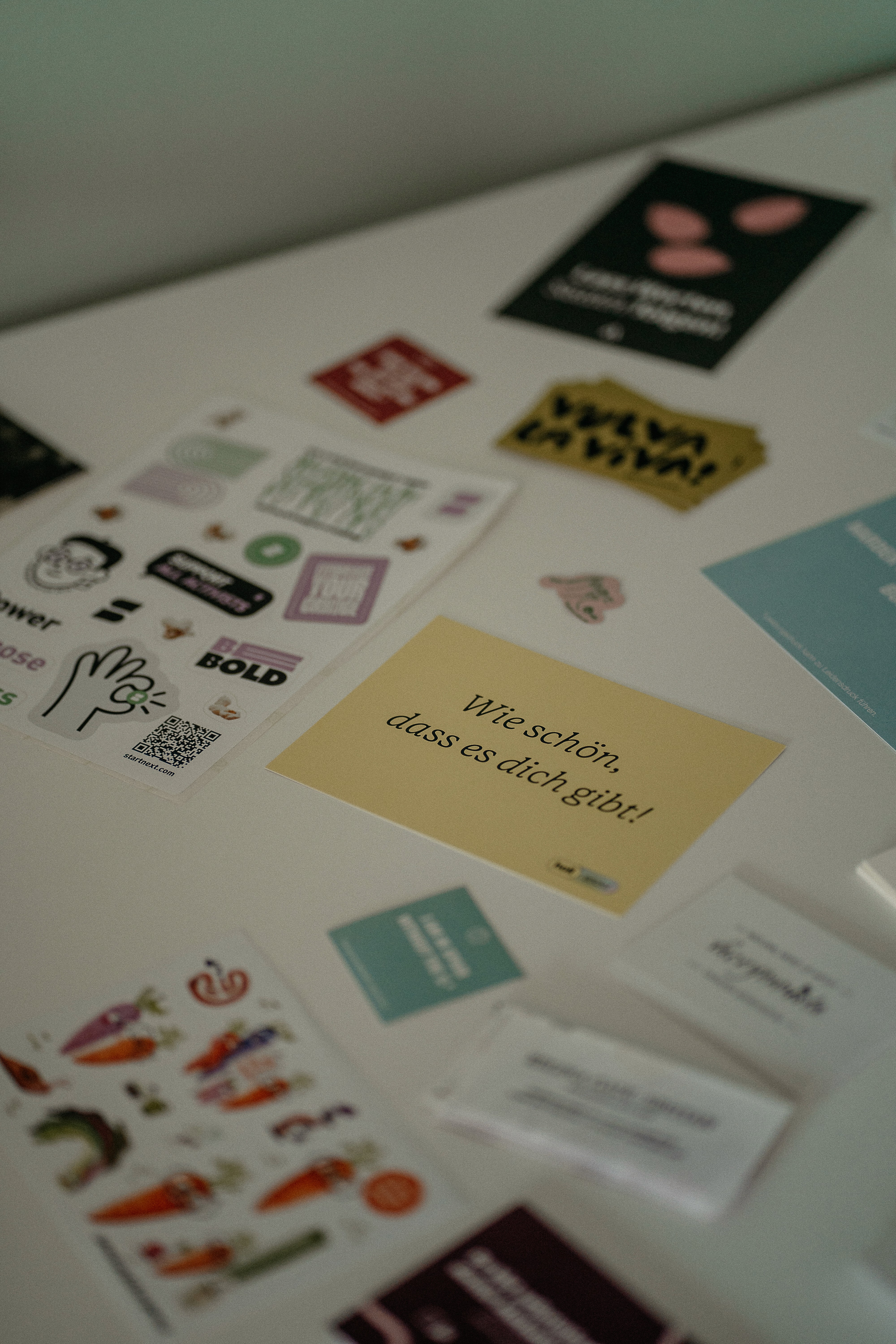 Auf einem Tisch liegen verschiedene Aufkleber und Karten, darunter eine Karte mit dem Text 'Lass Worten Saaten folgen', ein gelber Aufkleber mit dem Text 'VULVA LA VIVA', sowie weitere bunte Aufkleber mit Illustrationen und Texten.