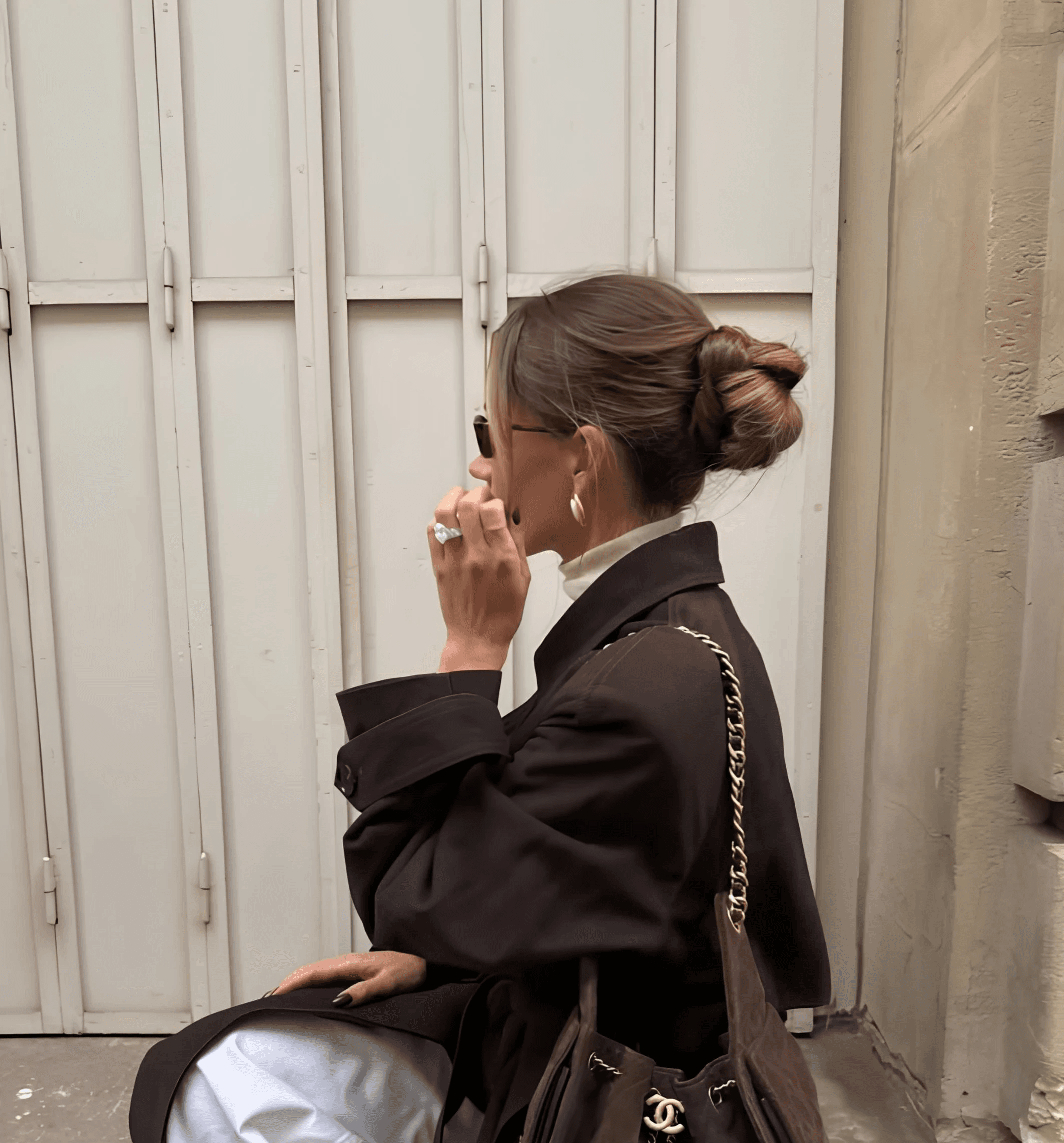 Woman seen in profile sitting on a stone ledge, wearing a chocolate brown trench coat over a cream turtleneck, white raw-hem jeans, sunglasses, gold hoop earrings and a low twisted bun, with a quilted brown suede Chanel bucket bag on a gold chain strap against a pale shuttered door.