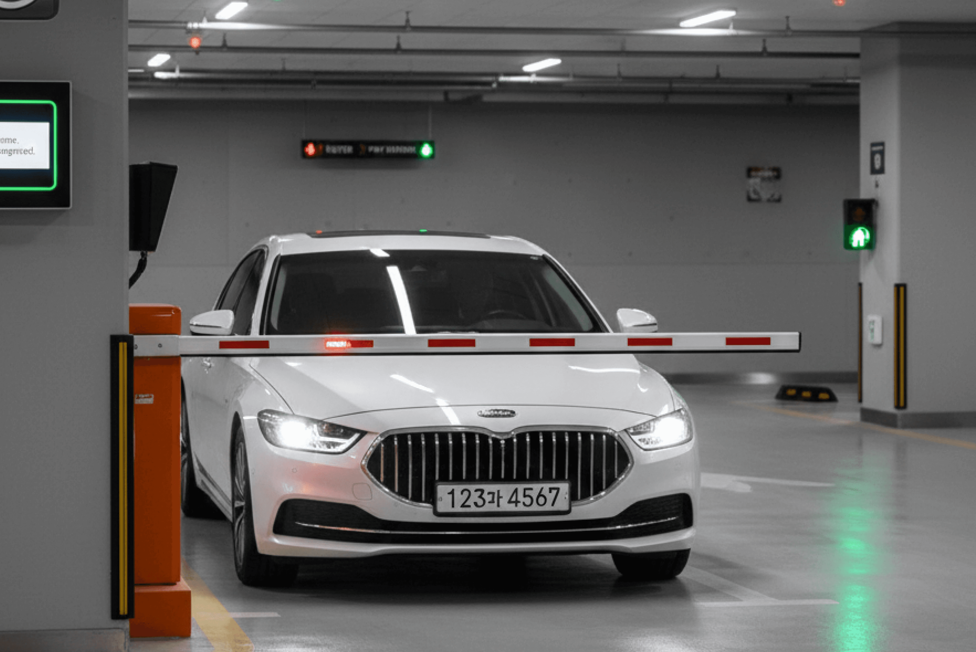 실내 주차장 입구에서 흰색 승용차(번호판 123과 4567)가 오렌지색 차단기 앞에 정차해 있으며, 좌측에 번호인식 카메라와 관제 디스플레이가 설치된 모습입니다.