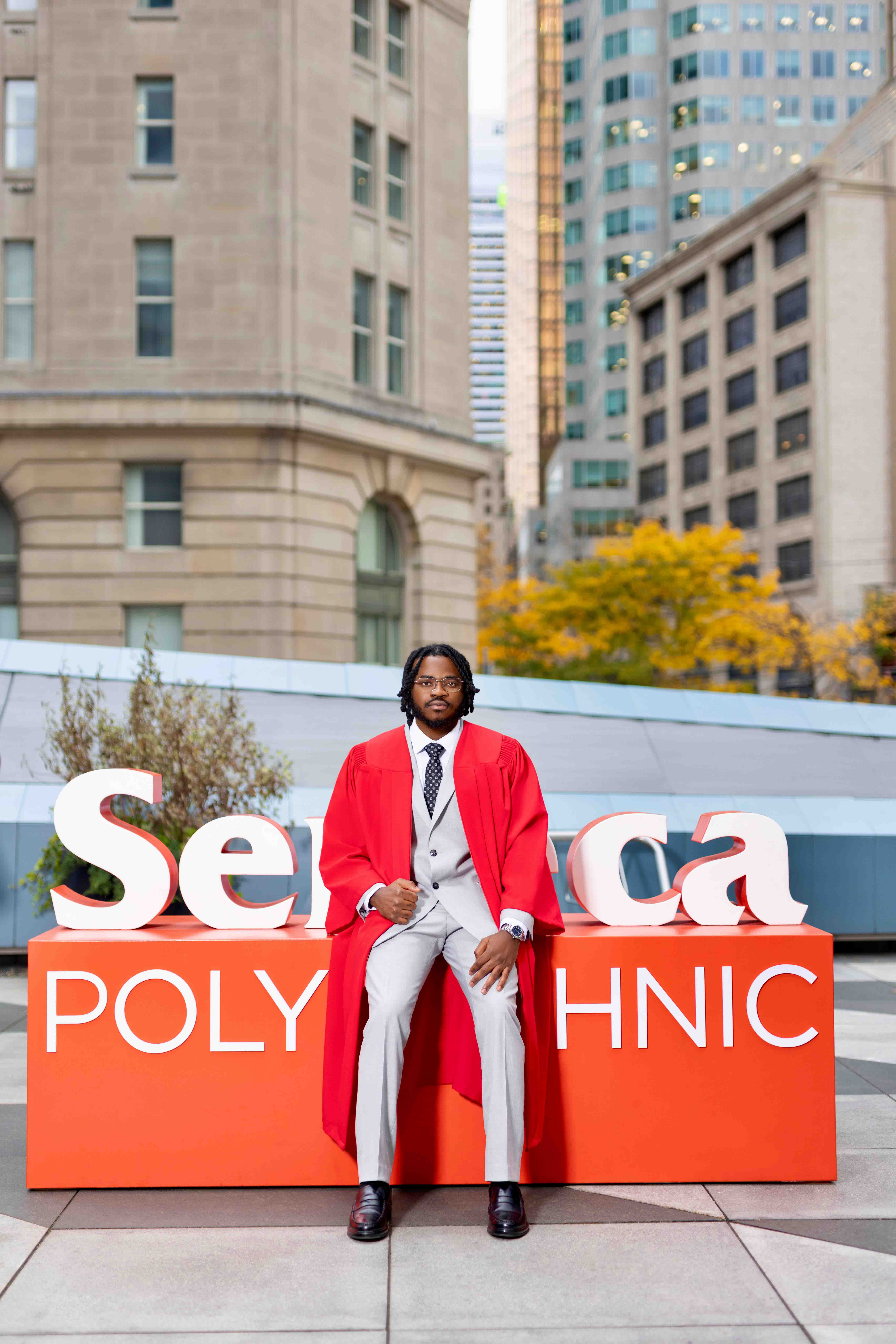 Graduate posing in a red robe in front of city buildings