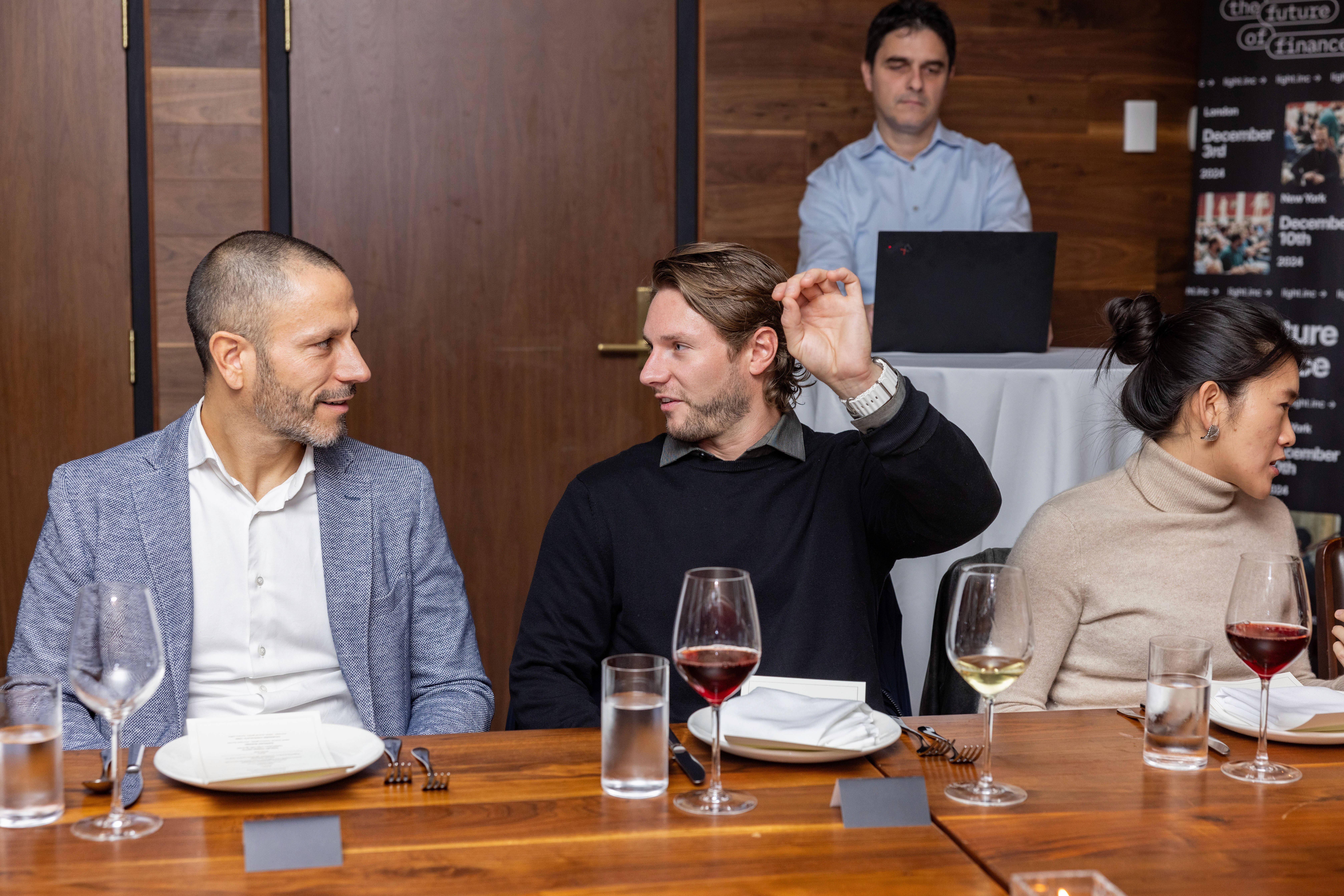 Two men and a woman sit at a dining table, engaged in conversation, with a server in the background.