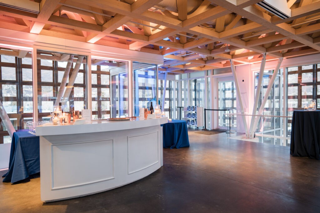 A sleek, white curved bar and blue cocktail tables set up for an evening reception at the Aspen Art Museum, showcasing professional corporate event photography. The shot highlights the venue's iconic wooden lattice ceiling, expansive glass walls, and warm ambient lighting.