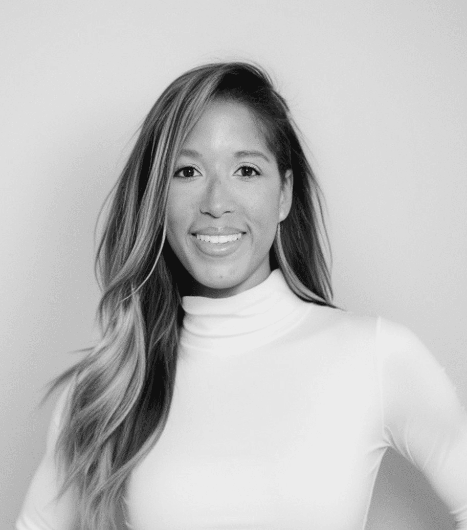 A smiling person with curly hair sits at a desk, looking at a laptop against a dark background.