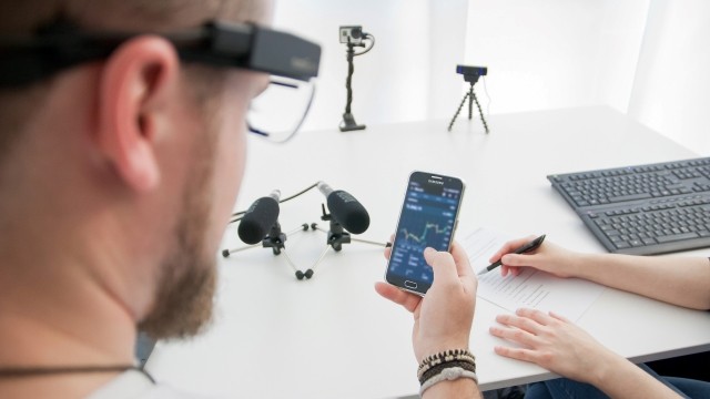 Person von hinten sitzt am Tisch und trägt Eye-Tracking-Brille, schaut auf Smartphone mit Diagrammen. 2 Hände von anderer Personen daneben liegen auf Tisch mit Stift. Auf Tisch sind Mikrofone, Kameras, Tastaturen.