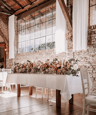 Lange Hochzeitstafel mit floraler Dekoration in rustikaler Location mit Backsteinwand in Essen