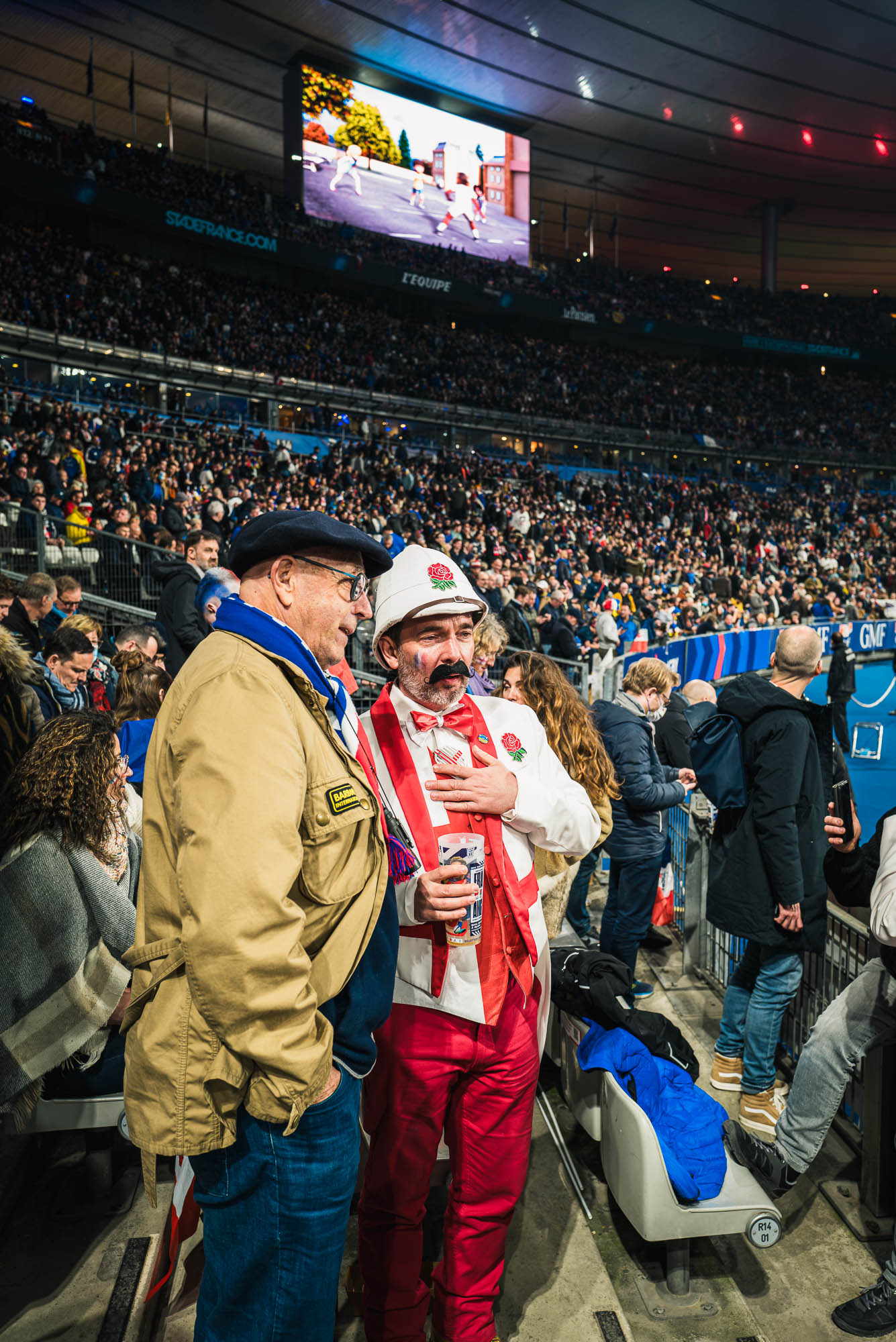 Un supporter français en béret et un supporter anglais en casque colonial, la rose d'Angleterre sur la poitrine, la main sur le cœur, discutent dans les gradins pendant la mi-temps