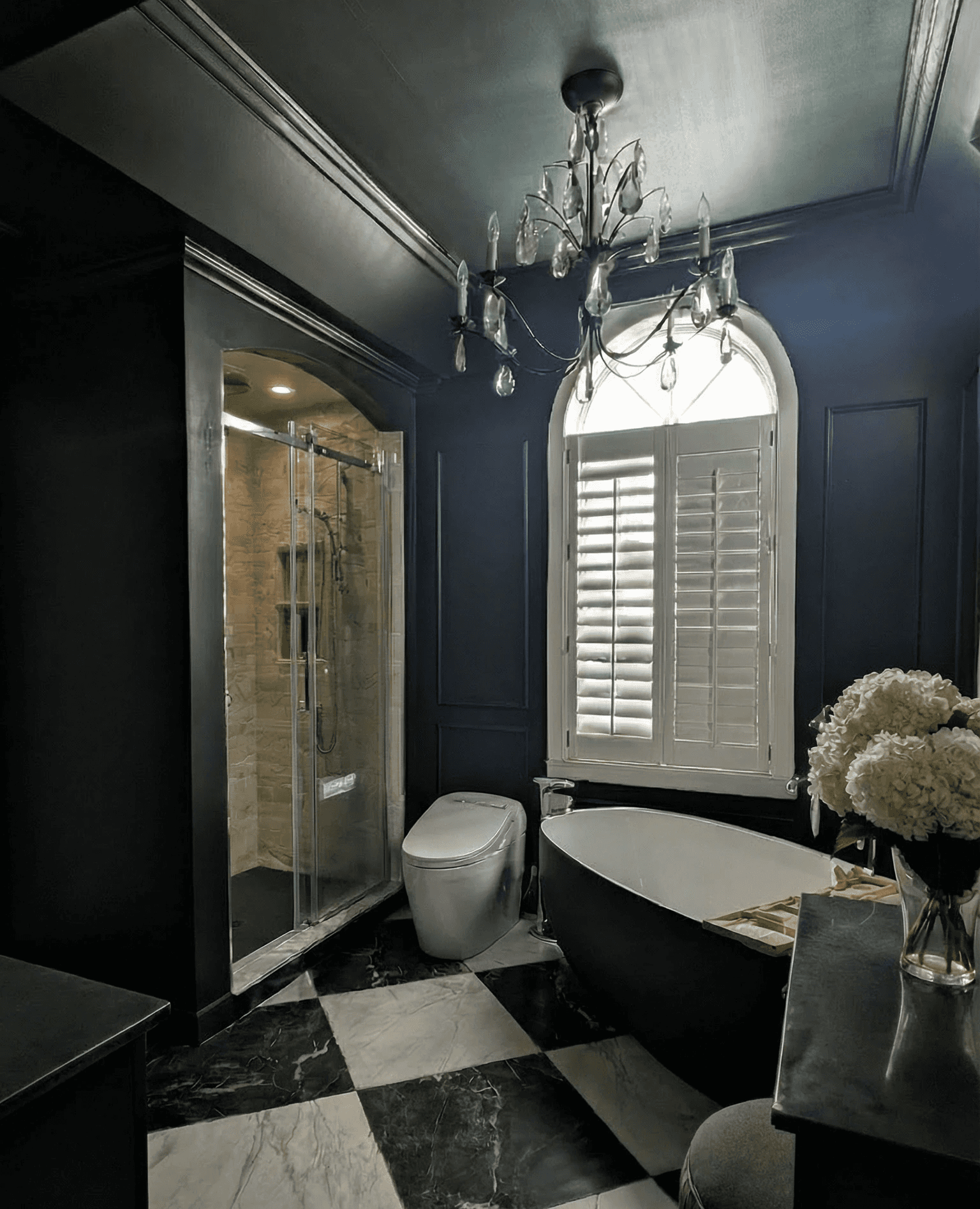 Salle de bains classique moody avec sol en damier noir et blanc veiné. Baignoire noire avec robinetterie autoportante chrome surmontée d'un magnifique lustre et d'une murale au plafond verte effet peinture à la chaux. Au plafond devant deux fenêtre en arche avec persiennes. Toilette blanche sans dossier. Coin maquilleuse avec meuble laqué brun avec un comptoir en dekton vert veiné. On aperçoit un banc vert en velour. On voit une douche dans le coin gauche, l'entrée de cette douche est en arche avec un porte de verre. La porcelaine de la douche est blanche et veinuré.  une base de douche noire. Deux alcôves supperposées son sur le mur intérieur de la douche.