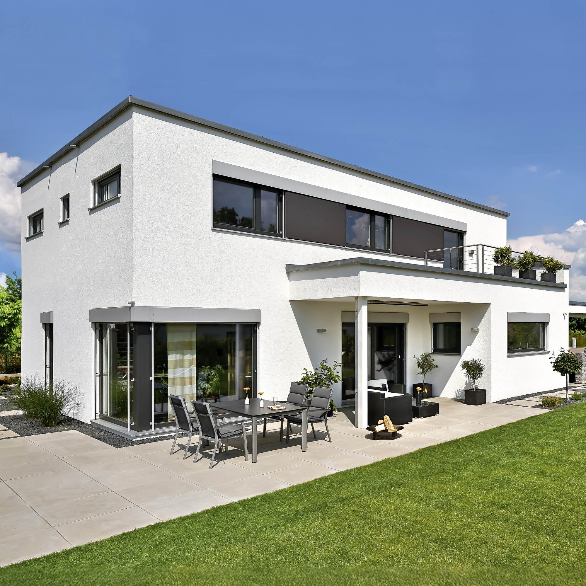 Modernes weißes zweistöckiges Haus mit großen Fenstern, Balkon und Terrasse mit Gartenmöbeln und Rasenfläche.