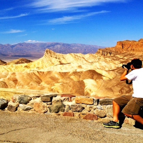 A person in a white t-shirt and shorts photographing a desert landscape with rock formations and mountains under a bright blue sky.