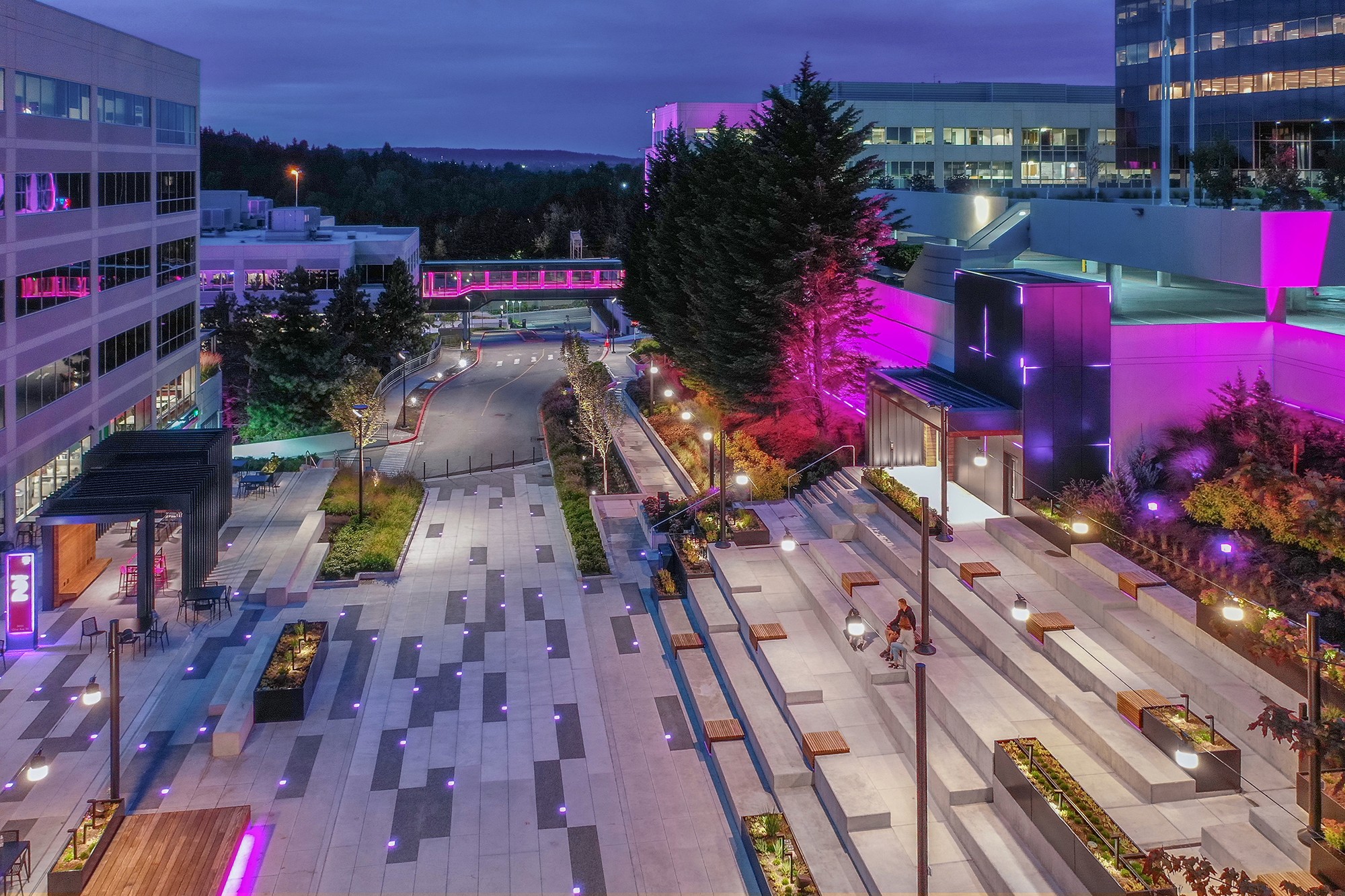 An aerial view of the vibrant new plaza at dusk, featuring glowing magenta digit lights embedded in sleek rectangular pavers and an accessible route winding up the amphitheater.