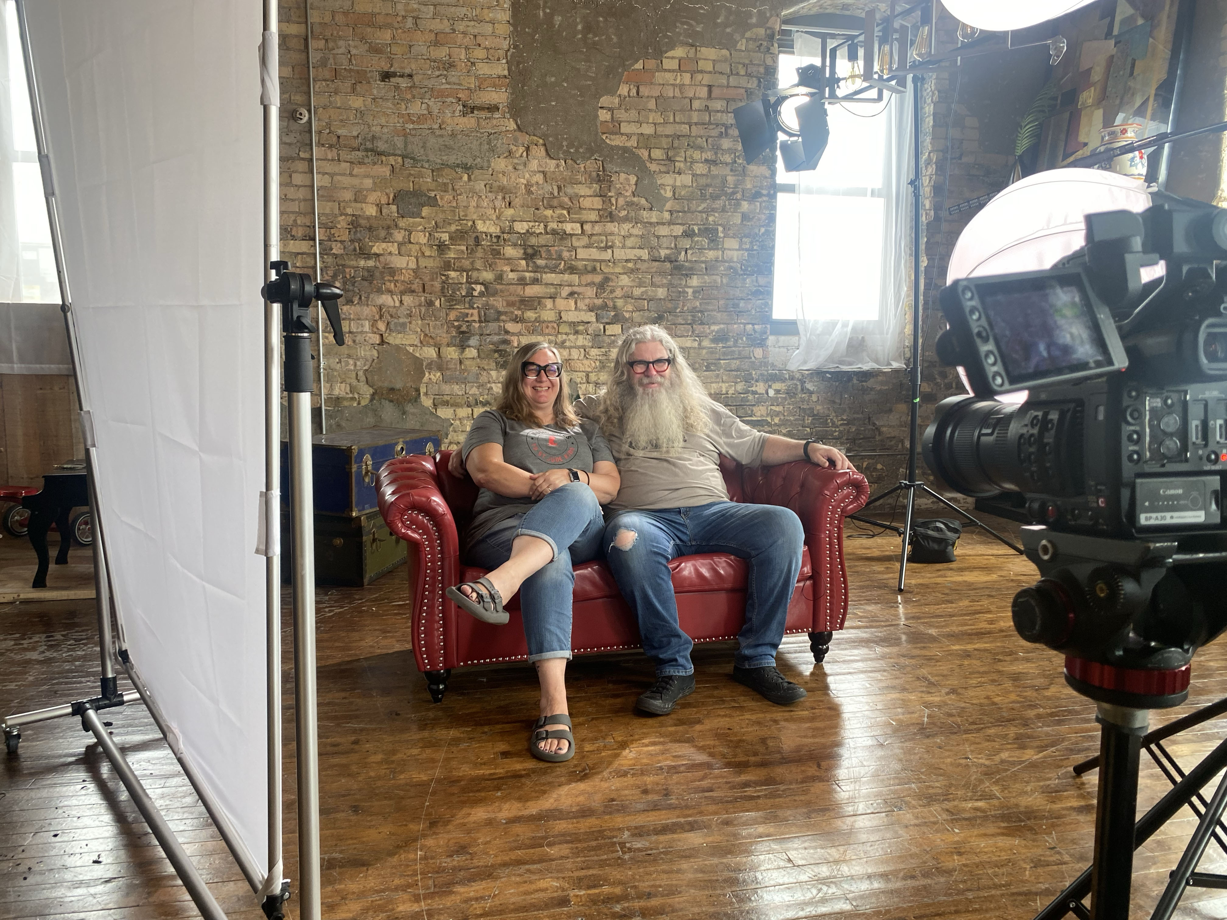 A man and woman sit on a red leather sofa inside a production studio with exposed brick walls and lighting gear.
