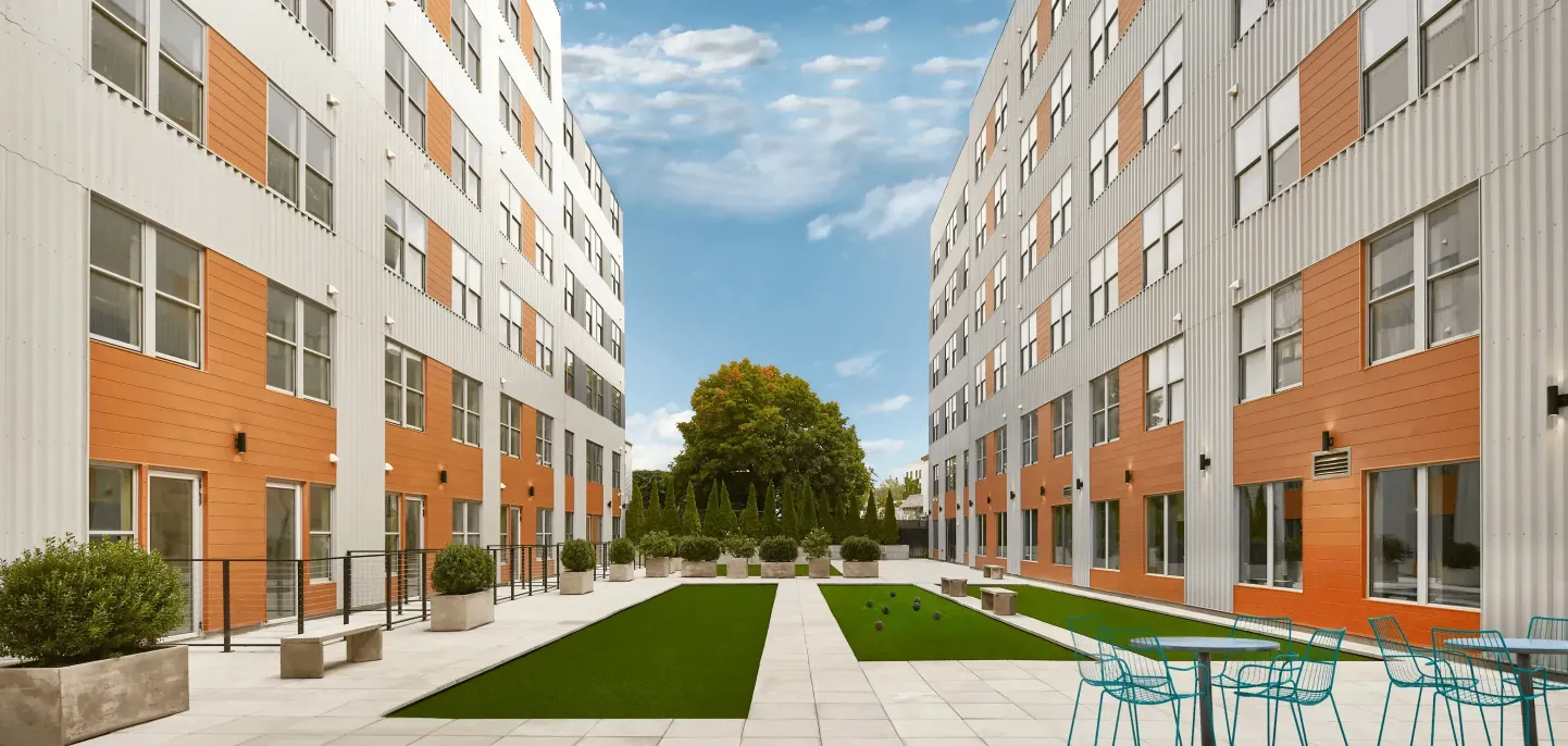 Modern courtyard flanked by buildings, featuring green grass and a tree, under a bright blue sky.