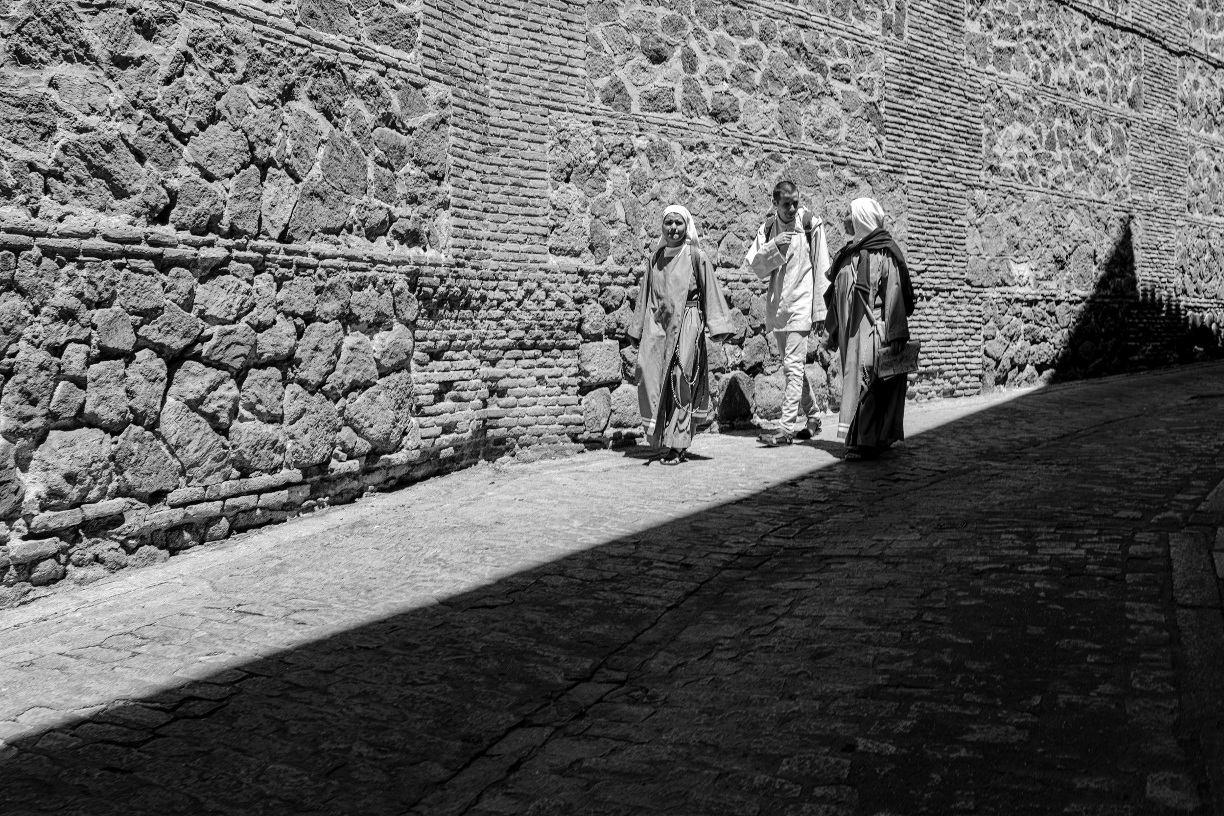 Three people walk along a cobblestone street beside a massive stone wall, sharply divided by a diagonal line of sunlight and shadow at midday.