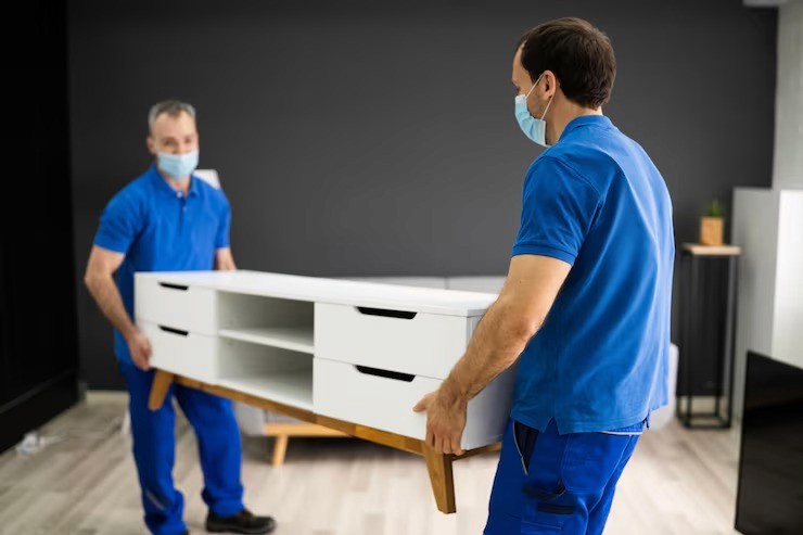 Two movers wearing masks carry a white entertainment unit indoors. Focus on movers and the furniture.