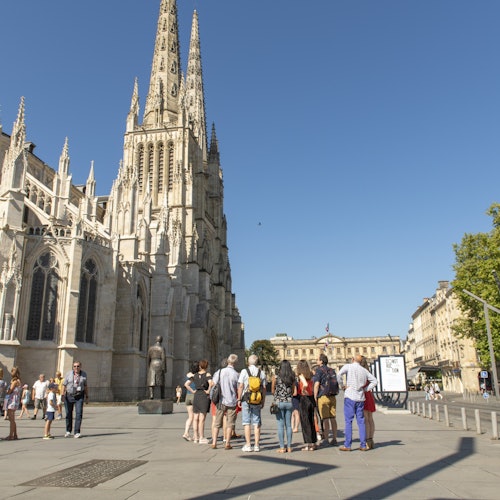 A group of people stands near a large, ornate cathedral with tall spires on a clear day, with buildings and trees in the background.