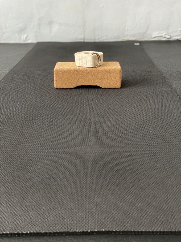 A beige woven strap rests on a cork yoga block, neatly placed on a dark textured yoga mat against a light gray background.