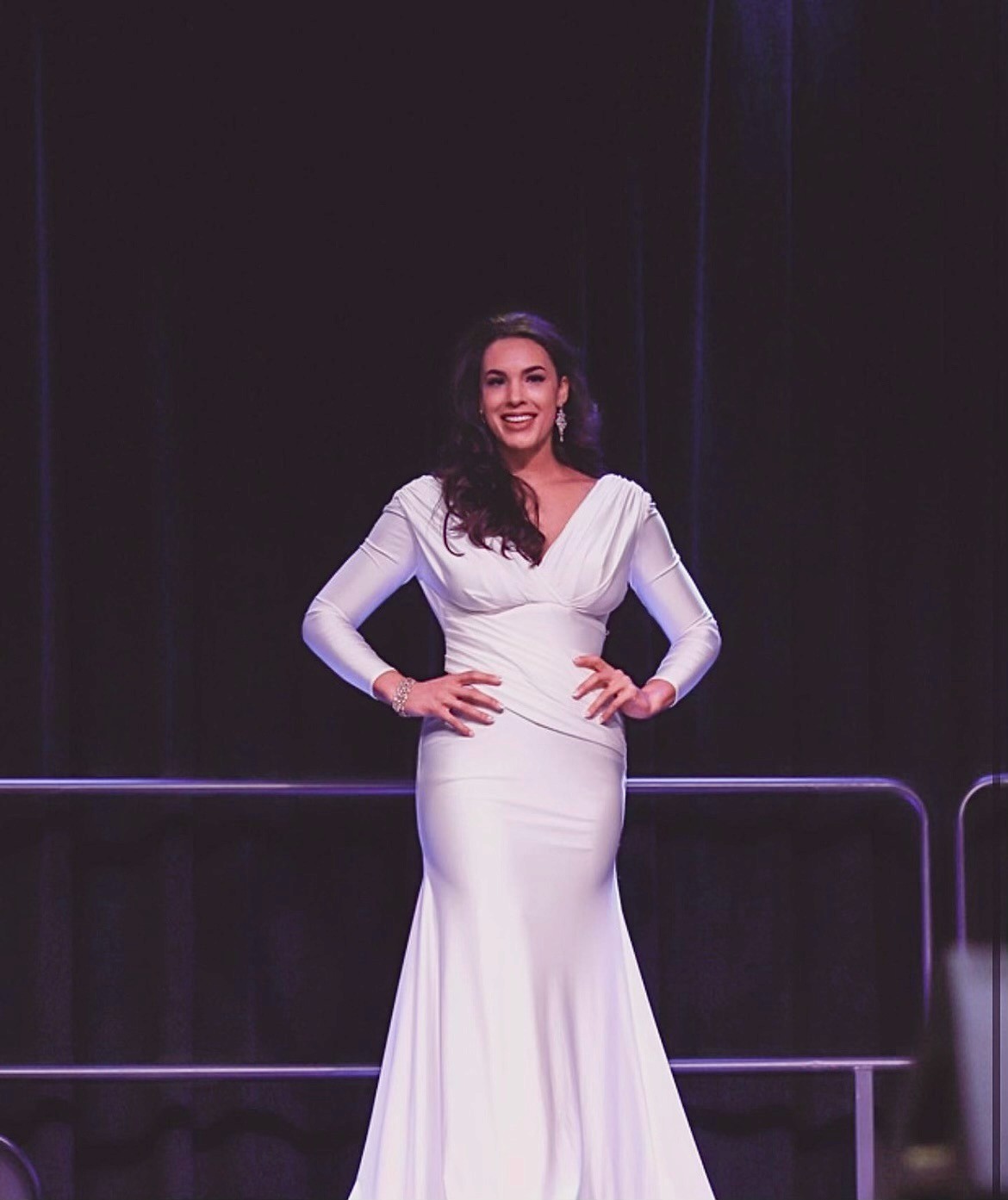 Elegant woman in a long white gown stands confidently on stage with hands on hips, smiling. Dark background adds focus and drama to her pose.