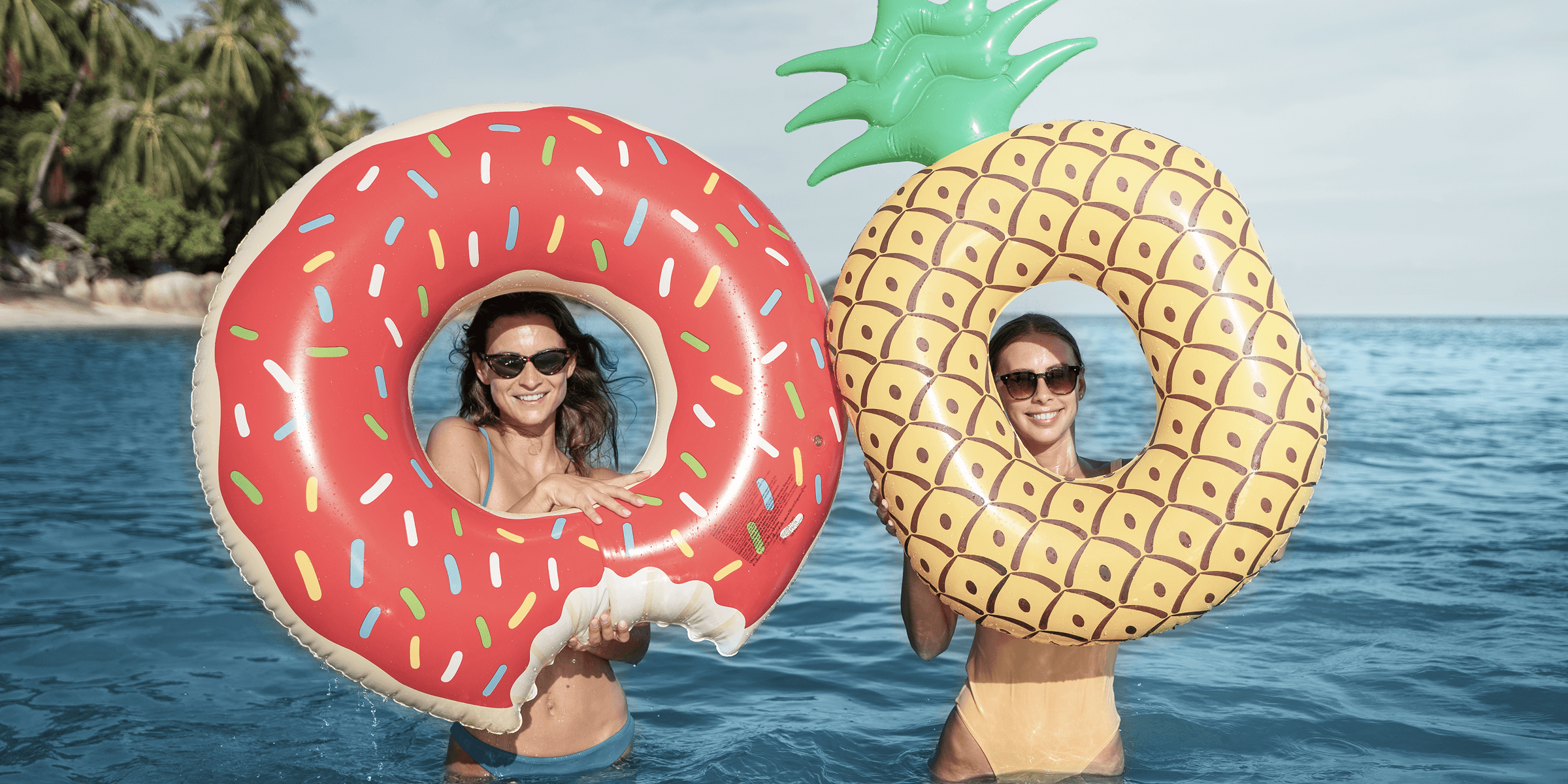 Two women smiling through donut and pineapple pool floats in the ocean.