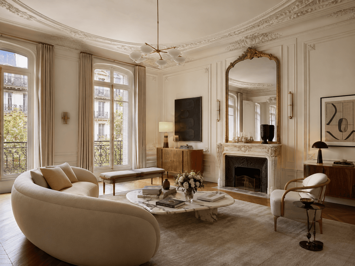 Elegant Paris Haussmann apartment living room with ornate moldings, tall French windows, marble fireplace, gilded mirror, and curved cream sofa.