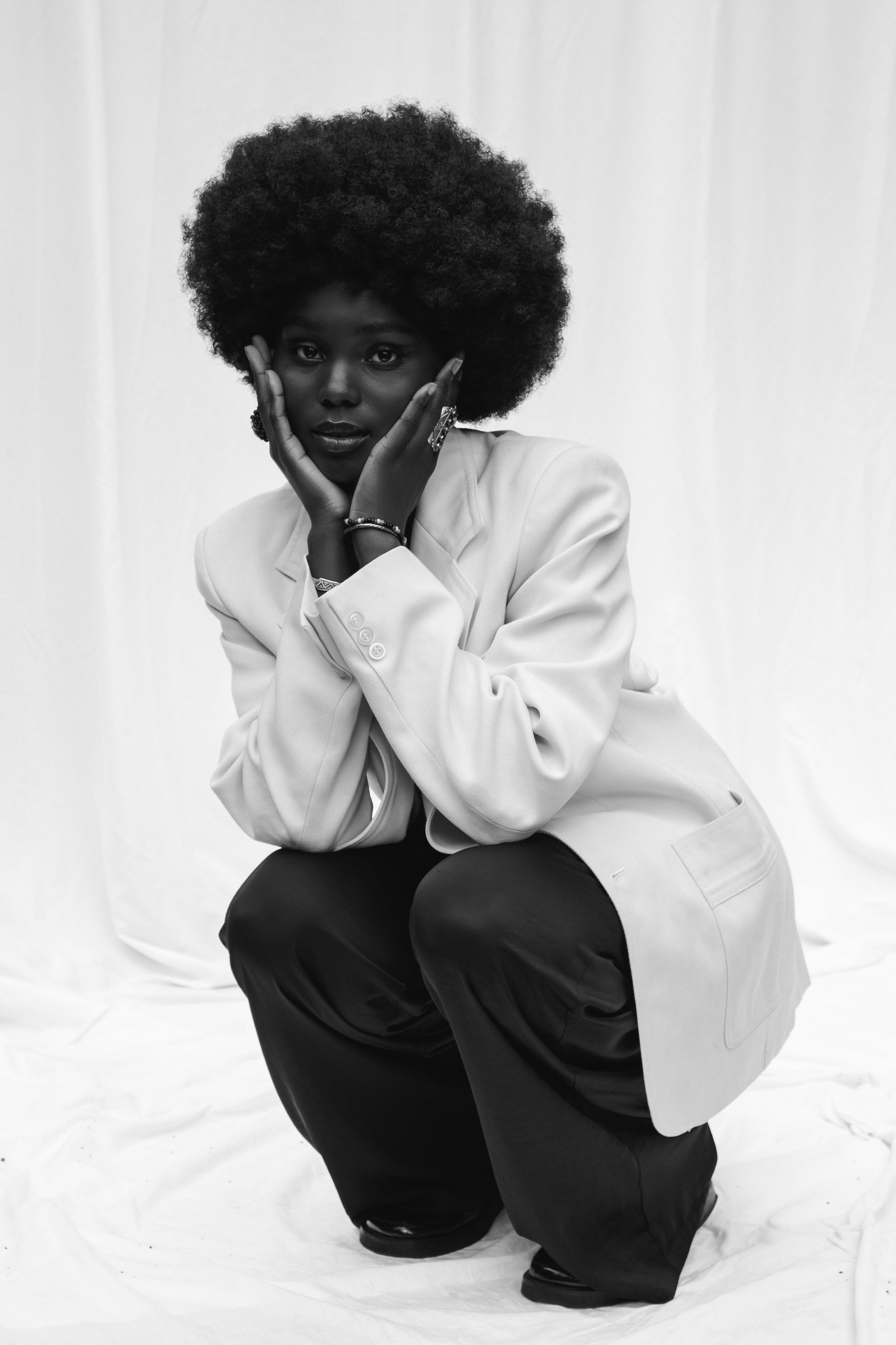 A person with an afro hairstyle crouches down, wearing a white shirt and dark pants, in a minimalist setting.