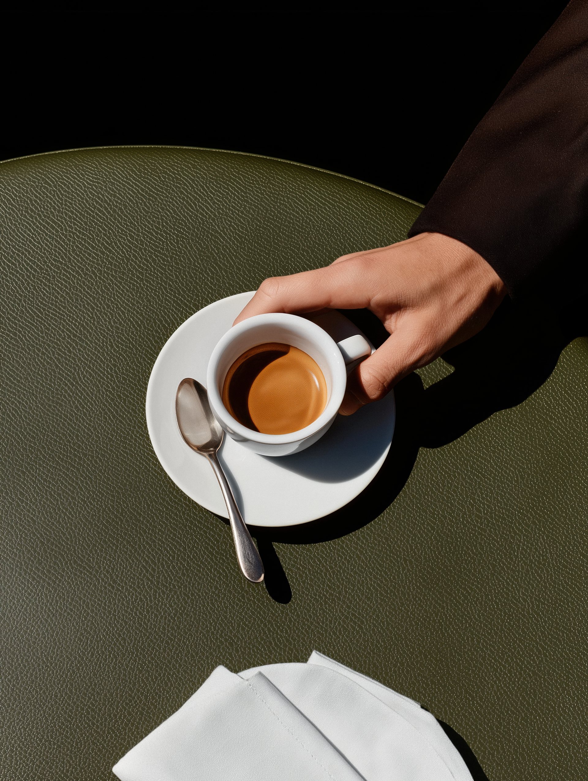 Hand lifting a white espresso cup from its saucer on a green leather table
